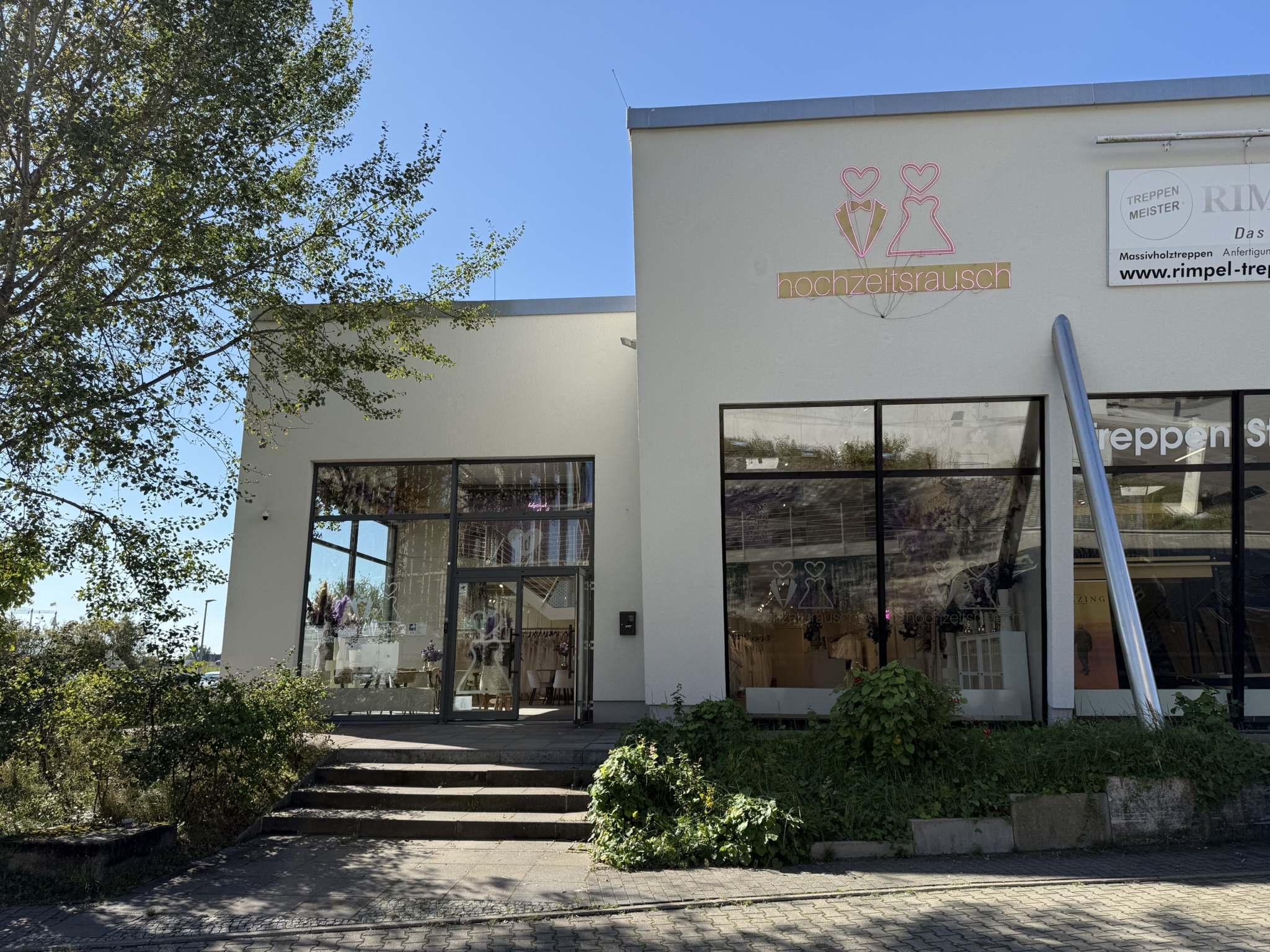 Das Brautmodengeschäft hochzeitsrausch Brautmoden Berlin Schönefeld verfügt über große Schaufenster, Schaufensterpuppen in Brautkleidern und ein Logo mit zwei Herzen und Hochzeitskleidern über dem Eingang. Begrünung und Stufen vervollständigen die einladende Fassade.