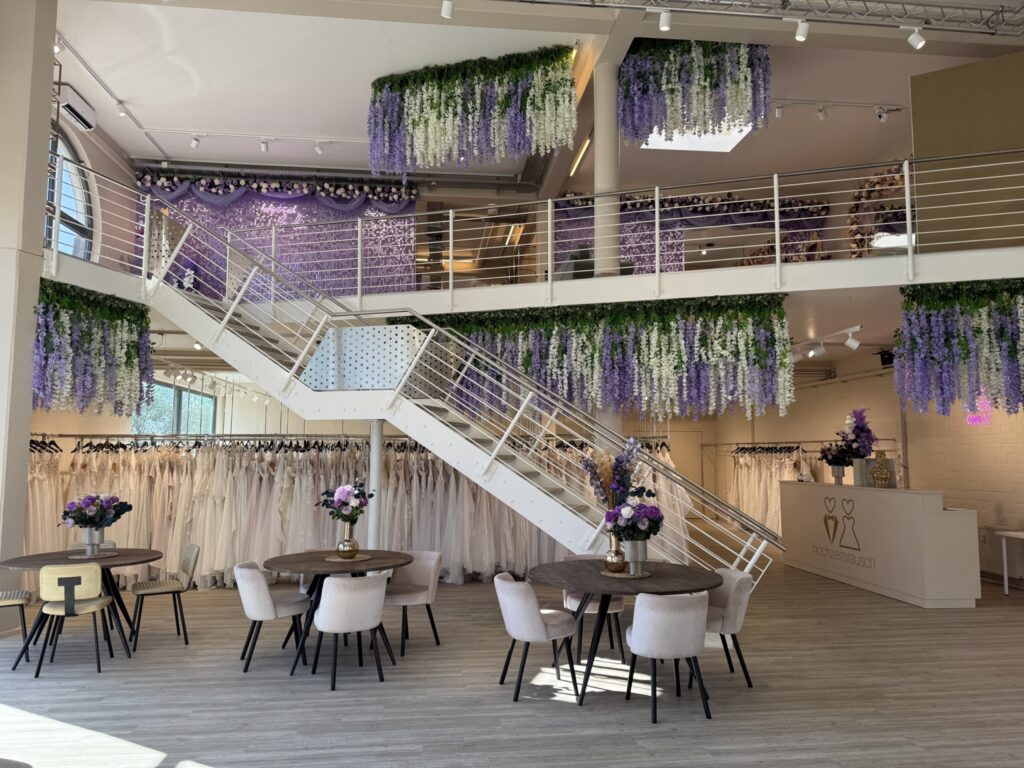 Ein geräumiges Brautmodengeschäft mit modernem Dekor, Blumenarrangements auf den Tischen, Regalen mit weißen Brautkleidern und hängenden lila und weißen Blumengirlanden. Natürliches Licht füllt den Raum im hochzeitsrausch Brautmoden Berlin Schönefeld.