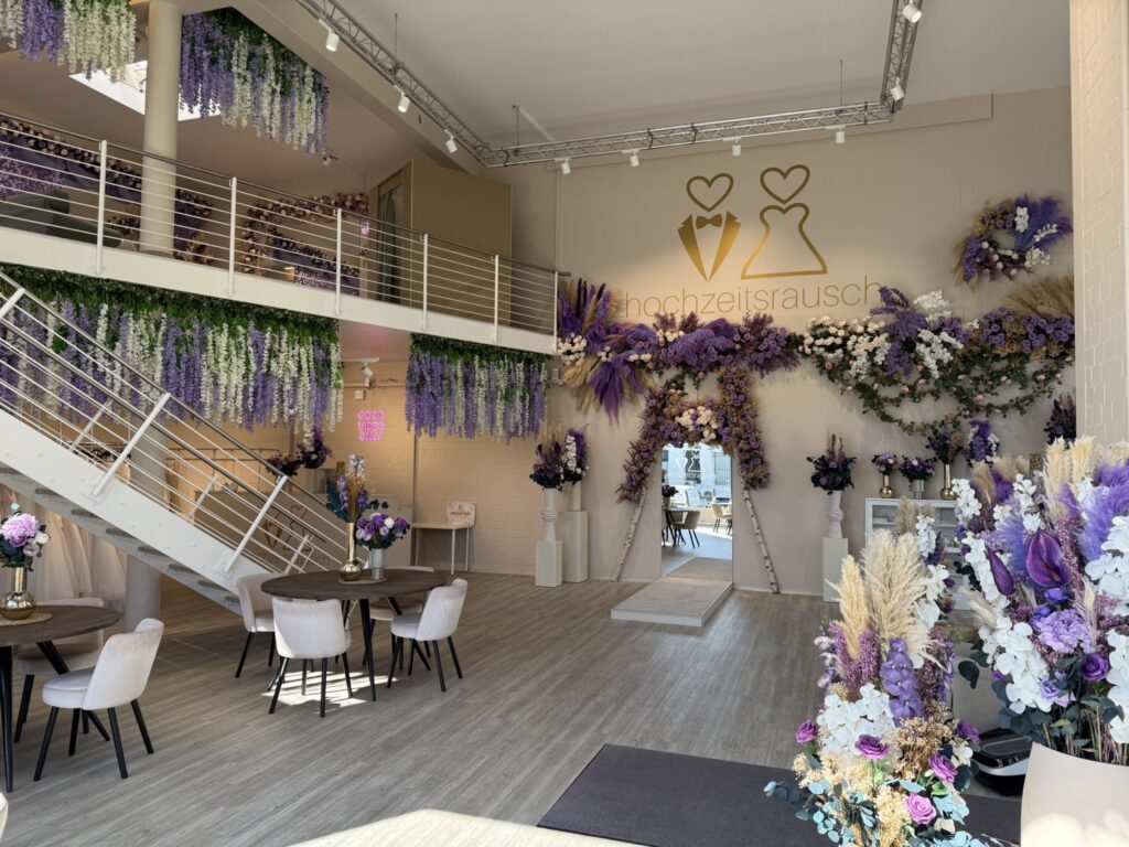 Ein moderner Veranstaltungsraum, dekoriert mit lila, weißen und rosafarbenen Blumen, eleganten Sitzgelegenheiten und einem Blumentorbogen. An der Wand prangt ein großes Hochzeitslogo und der Schriftzug hochzeitsrausch Brautmoden Berlin Schönefeld. Licht durchflutet den großzügigen Raum.
