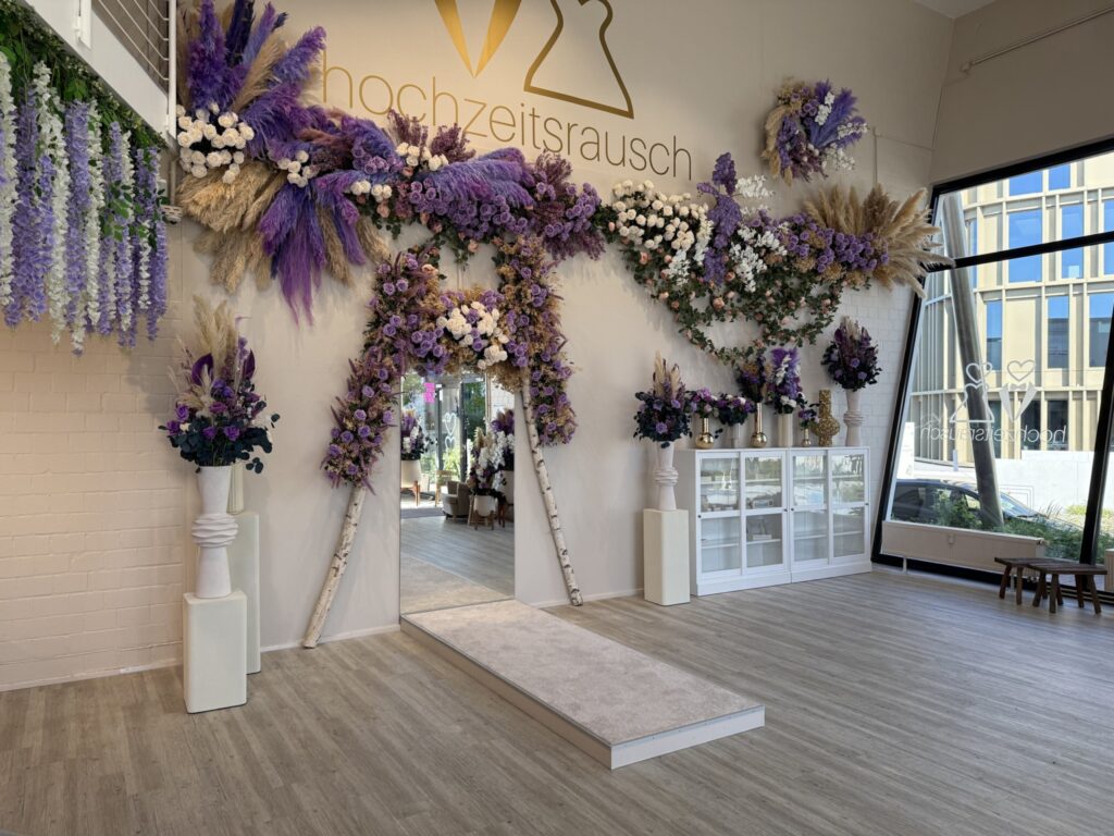 Ein Hochzeitsschauraum mit einem Blumenbogen aus lila, weißen und beigen Blumen, eleganten weißen Vasen, einem weißen Laufstegteppich und Hochzeitsrausch Brautmoden Berlin Schönefeld an der Wand. Große Fenster lassen natürliches Licht herein.