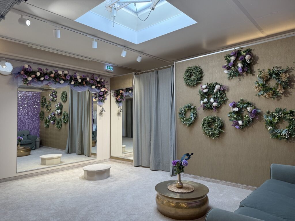 Eine stilvolle Umkleidekabine mit beigen Wänden, grauen Vorhängen, einem Oberlicht, Blumenkränzen an der Wand, einem großen Spiegel und einem goldenen Tisch mit einer Vase mit lila Blumen. Der Raum ist hell und elegant eingerichtet.