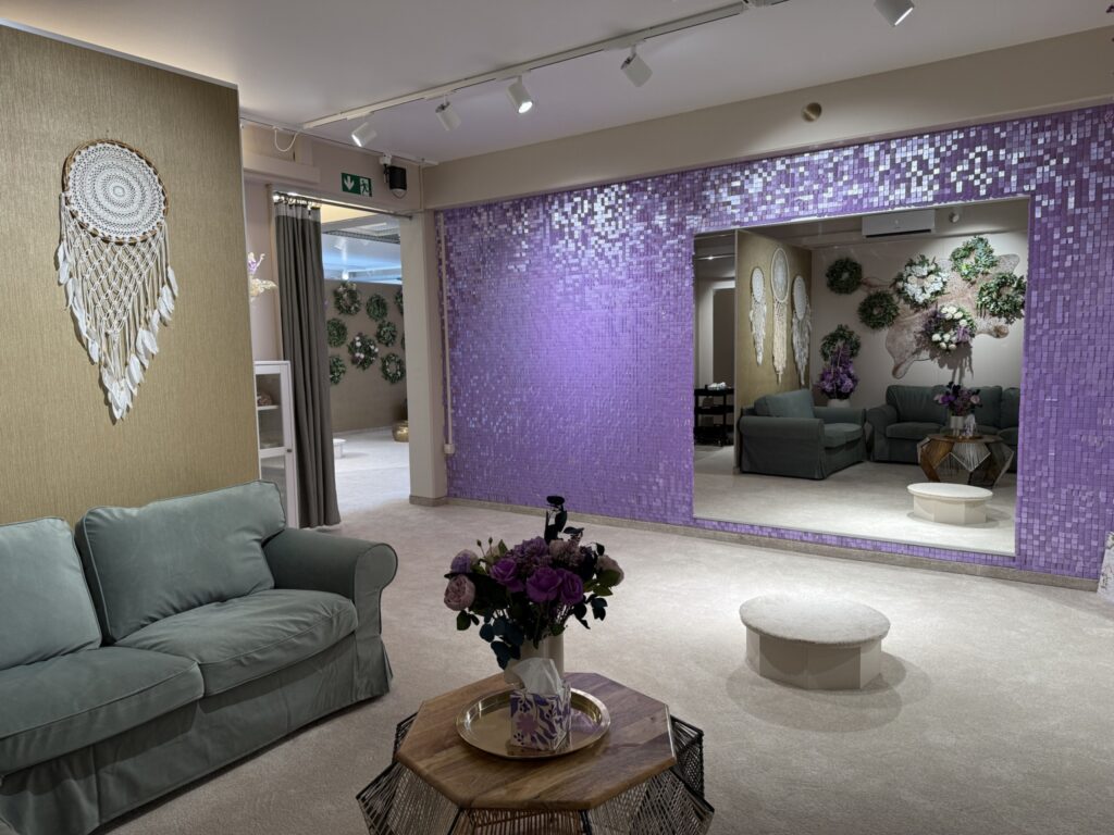 Ein modernes, sanft beleuchtetes Zimmer im hochzeitsrausch Brautmoden Berlin Schönefeld ist mit einem grünen Sofa, einem runden Tisch mit Blumenarrangement, einem großen lila gekachelten Spiegel, Traumfängern an der Wand und dekorativen Kränzen und Blumen ausgestattet.