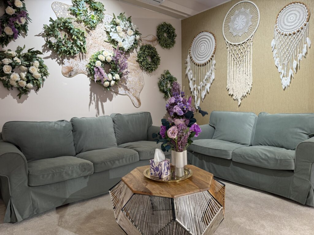 Ein gemütliches Wohnzimmer mit zwei grauen Sofas, einem geometrischen Couchtisch aus Holz mit einer Vase mit lila Blumen, Blumenkränzen an der Wand und großen dekorativen Traumfängern. Die Einrichtung ist weich und einladend.