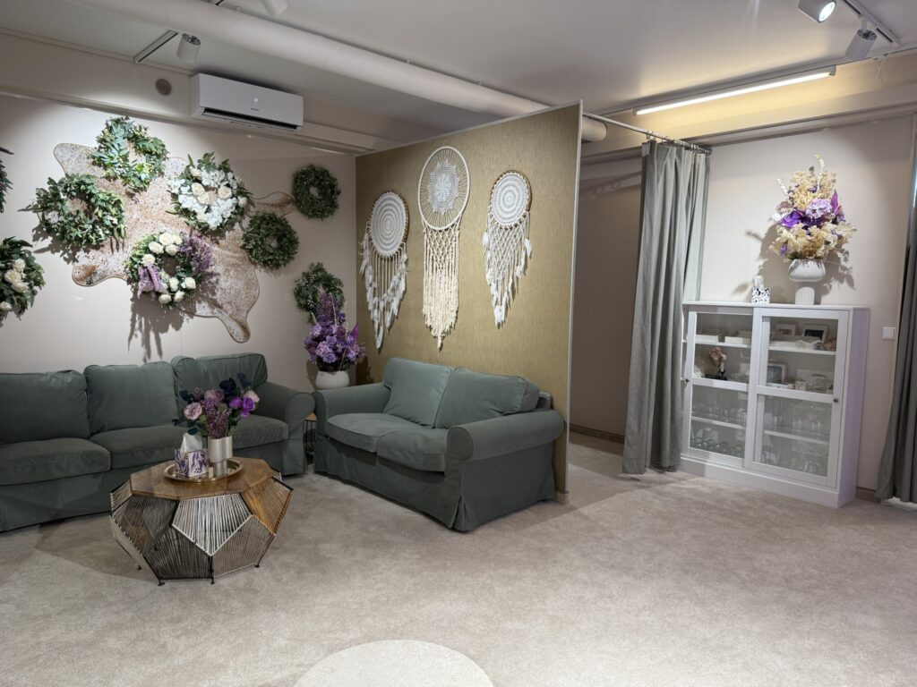 Ein gemütliches Wohnzimmer mit salbeigrünen Sofas, einem geometrischen Couchtisch aus Holz, Blumenarrangements, an der Wand befestigten Kränzen, großen Traumfängern und einem weißen Schrank mit Glastüren vor beigen Wänden und Teppichboden.