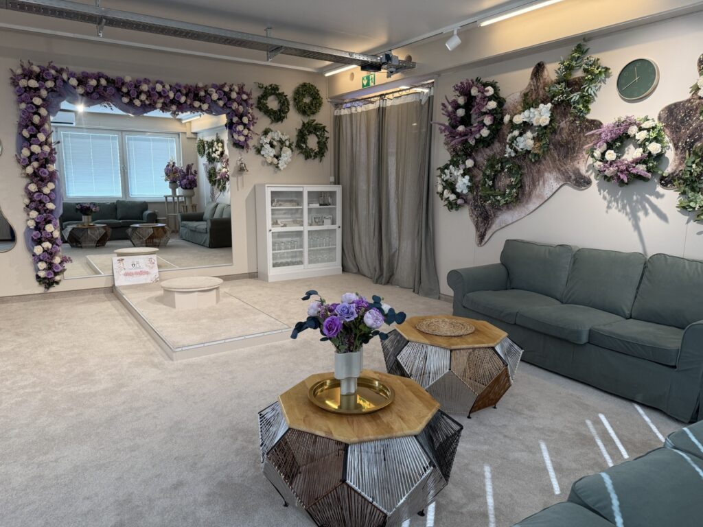 Ein gemütlicher, gut beleuchteter Raum mit grünen Sofas, Holztischen und Blumendekorationen in Lila, Weiß und Grün. An einer Wand befinden sich ein großer Spiegel und eine Vitrine, daneben Kränze und ein mit Blumen geschmückter Tierkopf.
