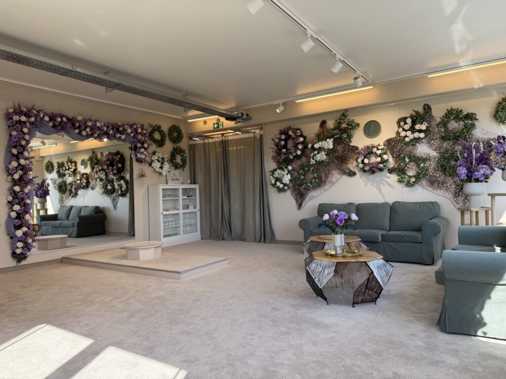 Eine helle, elegante Lounge im hochzeitsrausch Brautmoden Berlin Schönefeld ist mit grauen Sofas, einem geometrischen Couchtisch und lila und weißen Blumen geschmückt. Große Spiegel und sanfte Beleuchtung machen dieses Anprobezimmer einladend und gemütlich.