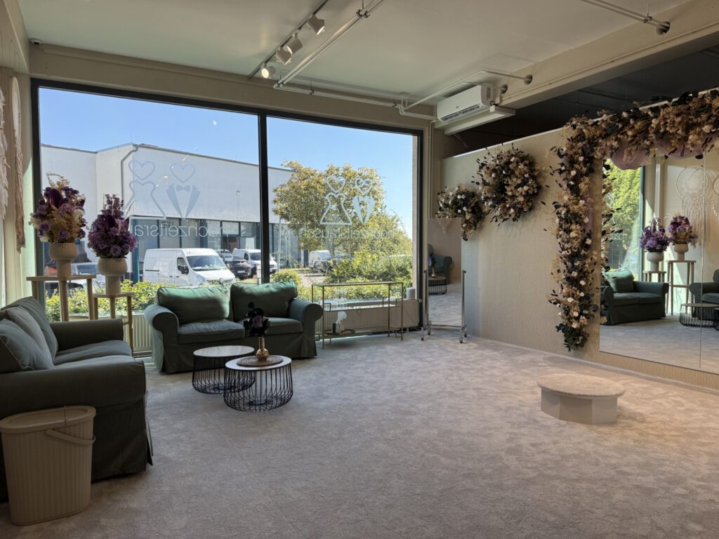 Ein helles, elegantes Anprobezimmer im hochzeitsrausch Brautmoden Berlin Schönefeld mit großen Fenstern, grünen Sofas, runden Couchtischen, einem geblümten Ganzkörperspiegel und einem kleinen runden Podest auf einem Teppichboden mit Blick auf Bäume und Gebäude draußen.