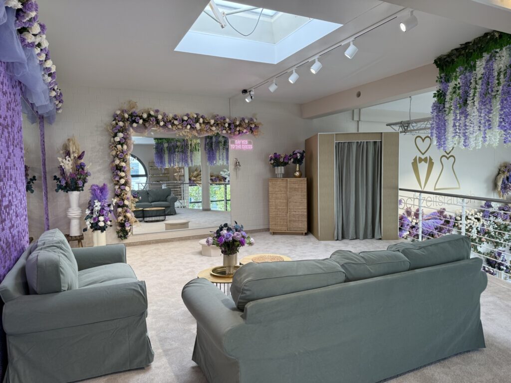 Ein geräumiges hochzeitsrausch Brautmoden Berlin Schönefeld Anprobezimmer mit pastellgrünen Sofas, Blumenarrangements in Lila und Weiß, einem großen, mit Blumen umrahmten Spiegel, einem runden goldenen Couchtisch, Oberlicht, Neon-Hochzeitsschild und einem vorgehangenen Anprobebereich.