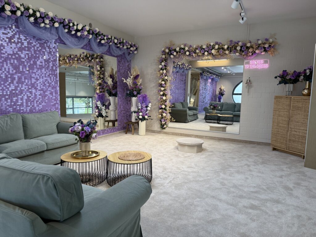 Die stilvolle Lounge im hochzeitsrausch Brautmoden Berlin Schönefeld ist mit hellgrünen Sofas, goldenen und schwarzen Couchtischen und großen, von Blumen eingerahmten Spiegeln ausgestattet. Lila Mosaikfliesen, Blumengirlanden und ein neonfarbenes "SAY YES TO THE DRESS"-Schild vervollständigen das Anprobezimmer.