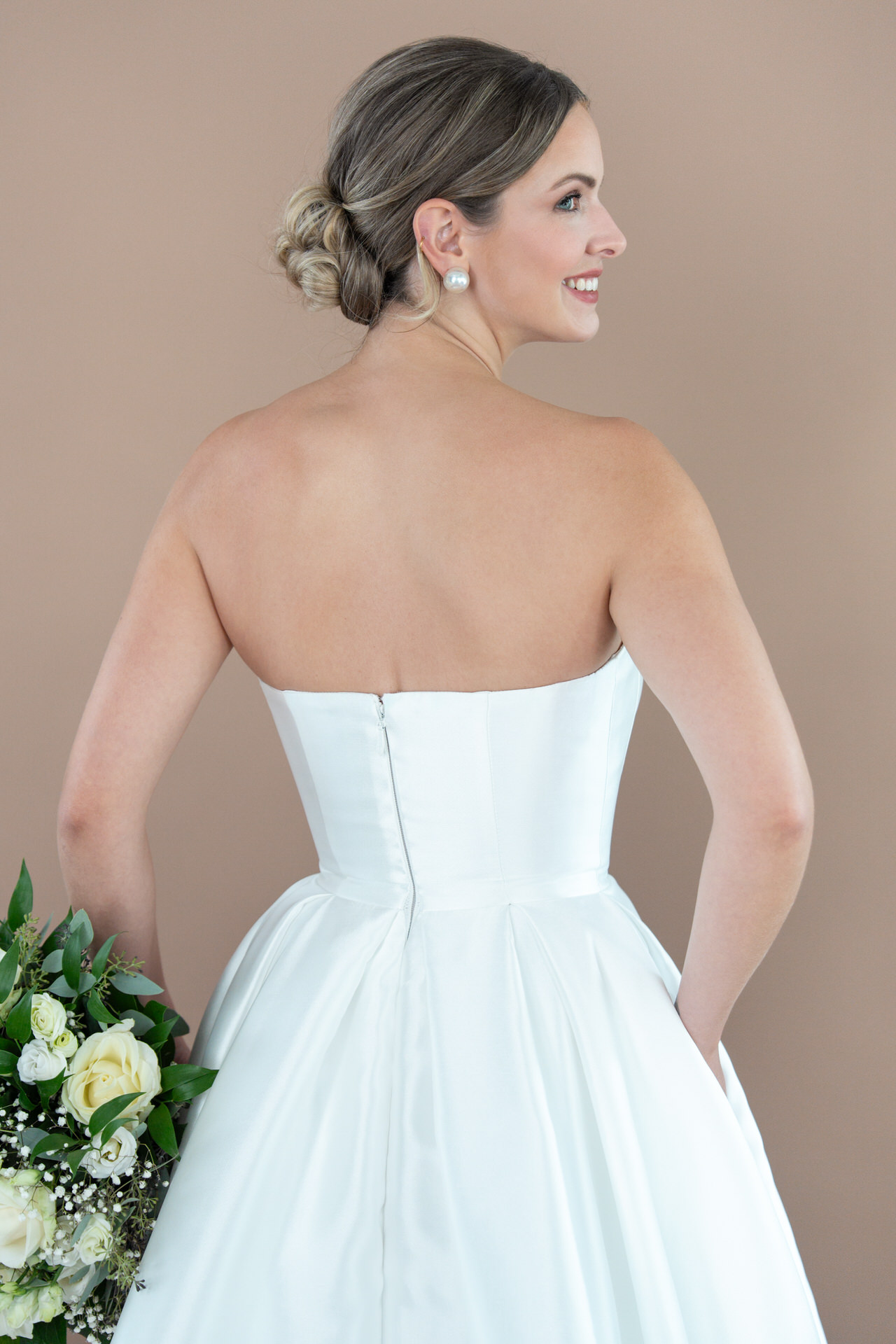 Eine Braut in einem trägerlosen weißen Bridonista-Kleid R2L26-C-A06 steht mit dem Rücken zur Kamera und hält einen Strauß weißer Rosen und Grünzeug in der Hand. Ihr Haar ist zu einem tiefen Dutt gebunden, während sie über ihre rechte Schulter lächelt.