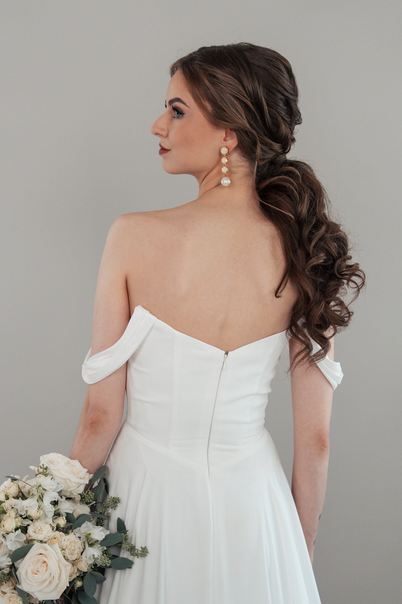 Das Modell S26-A01 zeigt eine Braut mit langen, gewellten braunen Haaren in einem lockeren Pferdeschwanz, die ein schulterfreies weißes Kleid trägt und weiße und cremefarbene Blumen in der Hand hält. Sie steht vor einem schlichten grauen Hintergrund und blickt leicht weg.