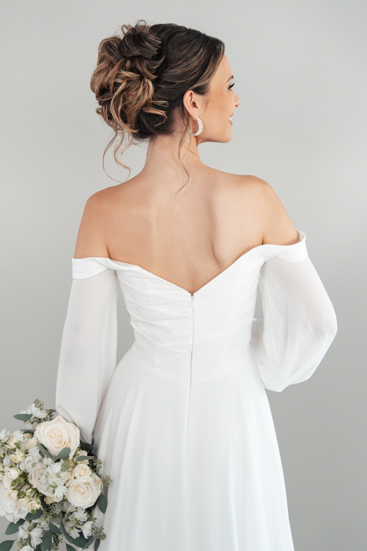 Eine Braut im schulterfreien weißen Hochzeitskleid S26-A02 steht mit dem Gesicht zur Wand und hält einen Strauß aus weißen Rosen und Grünzeug. Ihr Haar ist zu einer eleganten Hochsteckfrisur mit Ohrringen vor einem schlichten, hellen Hintergrund gestylt.