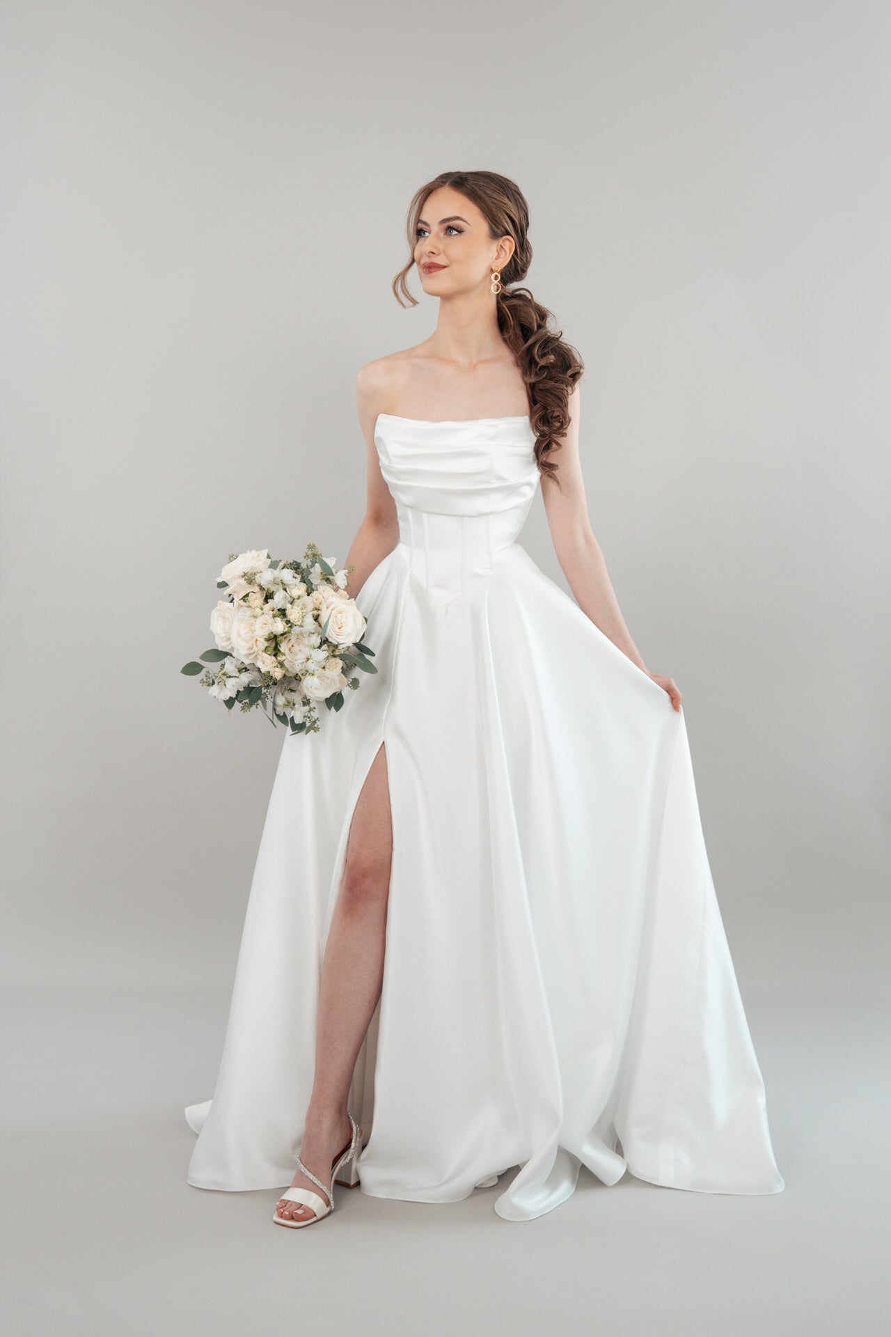 Eine Frau modelliert das S26-A06, ein trägerloses weißes Hochzeitskleid mit hohem Schlitz, und hält einen Strauß weißer Blumen und Grünzeug vor einem hellgrauen Hintergrund.