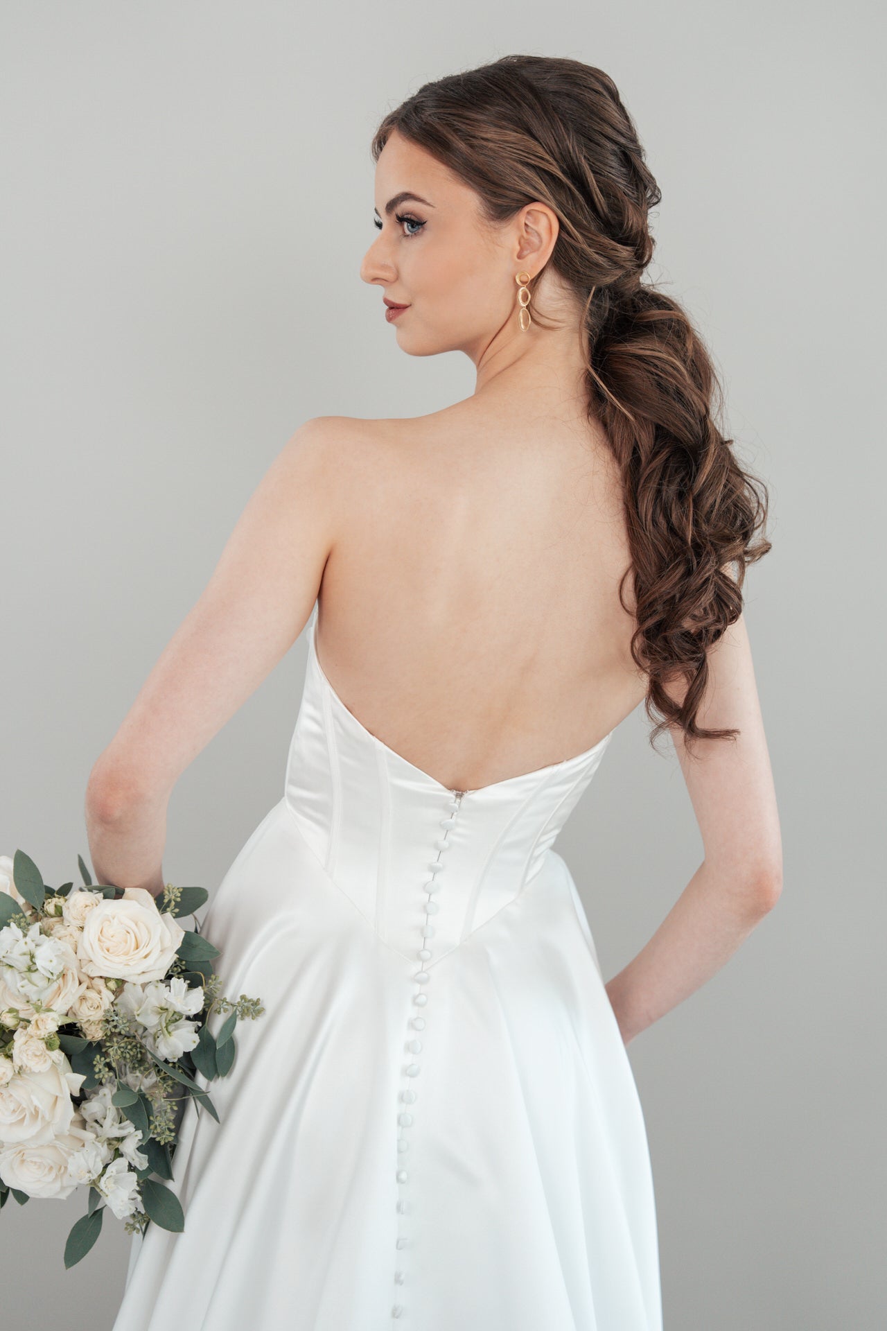 Eine Braut im trägerlosen weißen Hochzeitskleid S26-A06 steht mit dem Rücken zur Kamera und hält einen Strauß aus weißen Blumen und Grünzeug. Ihr langes braunes Haar ist in lockeren Wellen gestylt, dazu trägt sie goldene Tropfenohrringe.