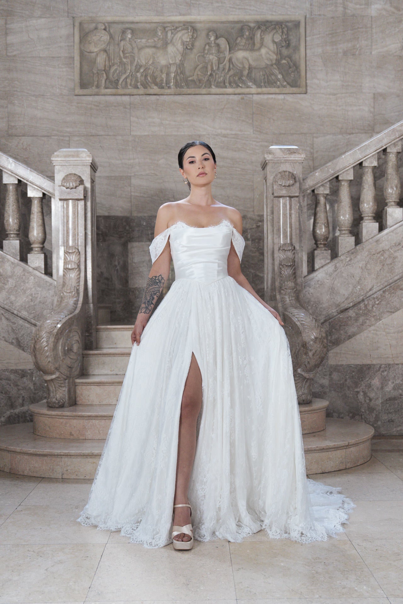 In dem schulterfreien weißen Hochzeitskleid S26-A07 mit oberschenkelhohem Schlitz steht eine Frau auf einer eleganten Steintreppe, eingerahmt von einem kunstvollen Geländer und einem geschnitzten Wandrelief hinter ihr.