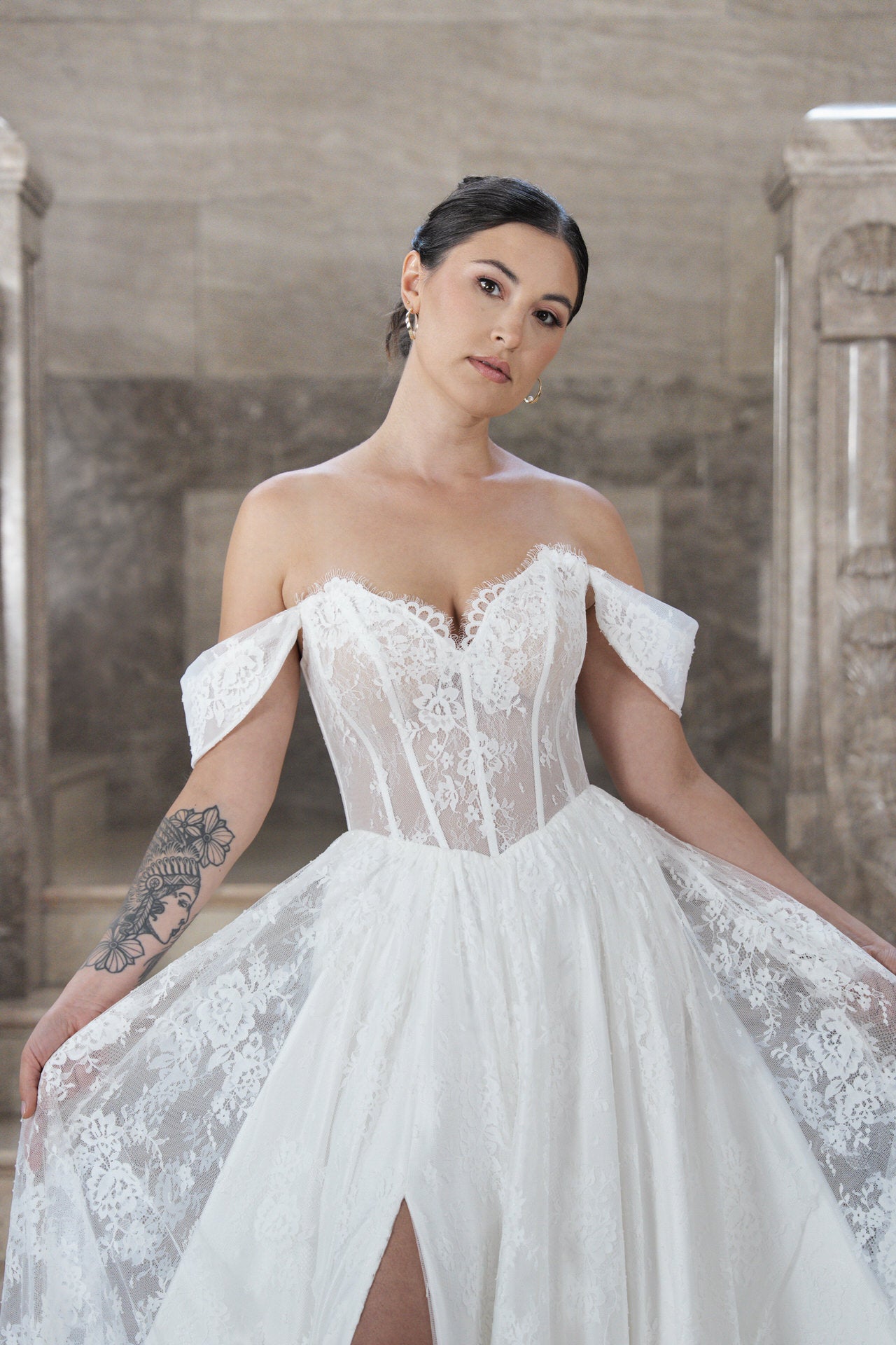 Eine Frau in dem schulterfreien Spitzenhochzeitskleid S26-A07 mit durchscheinendem Mieder posiert in einem Innenraum. Ihr dunkles Haar ist hochgesteckt, sie hat ein florales Armtattoo und steht vor einem Stein, auf dem sie ihr Kleid ausbreitet.