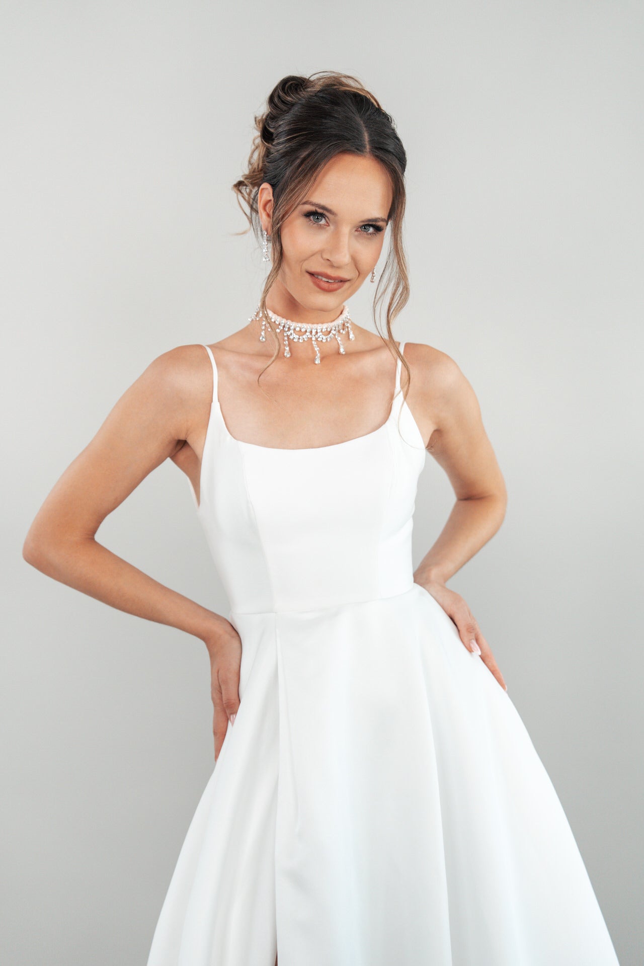 In dem Kleid S26-A08 posiert eine Frau mit den Händen in den Hüften; sie kombiniert den weißen, ärmellosen, ausgestellten Rock mit einem Halsband aus Perlen und Kristallen und trägt eine elegante Hochsteckfrisur vor einem hellgrauen Hintergrund.