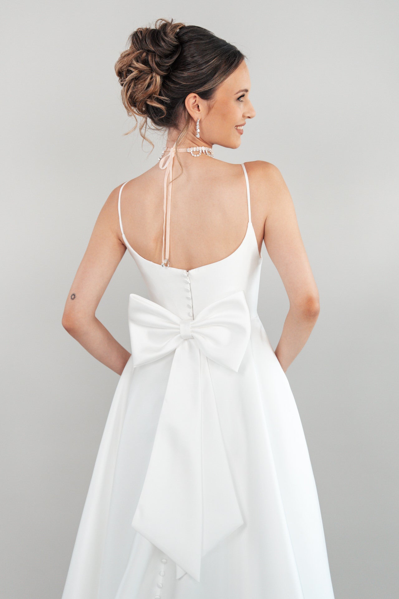 Eine Frau modelliert das weiße, ärmellose Kleid S26-A08 mit einer großen Schleife am Rücken. Ihre elegante Hochsteckfrisur und die zarten Ohrringe sind zu sehen, während sie sich vor einem schlichten, hellen Hintergrund abwendet.