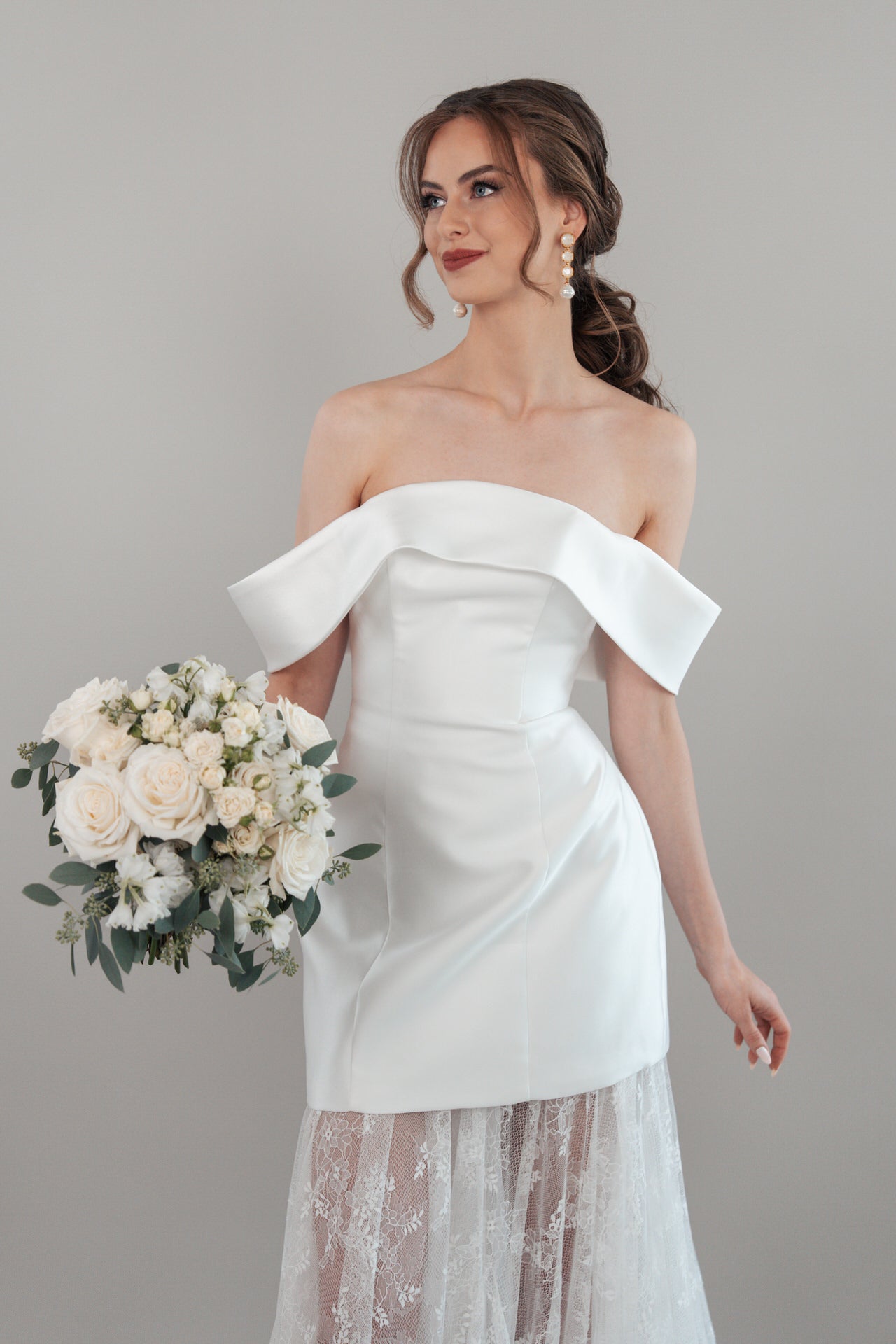 Eine Frau, die das schulterfreie weiße Brautkleid S26-A11 mit Spitzendetails trägt, hält einen Strauß weißer Rosen und Grünzeug vor einem schlichten hellgrauen Hintergrund.