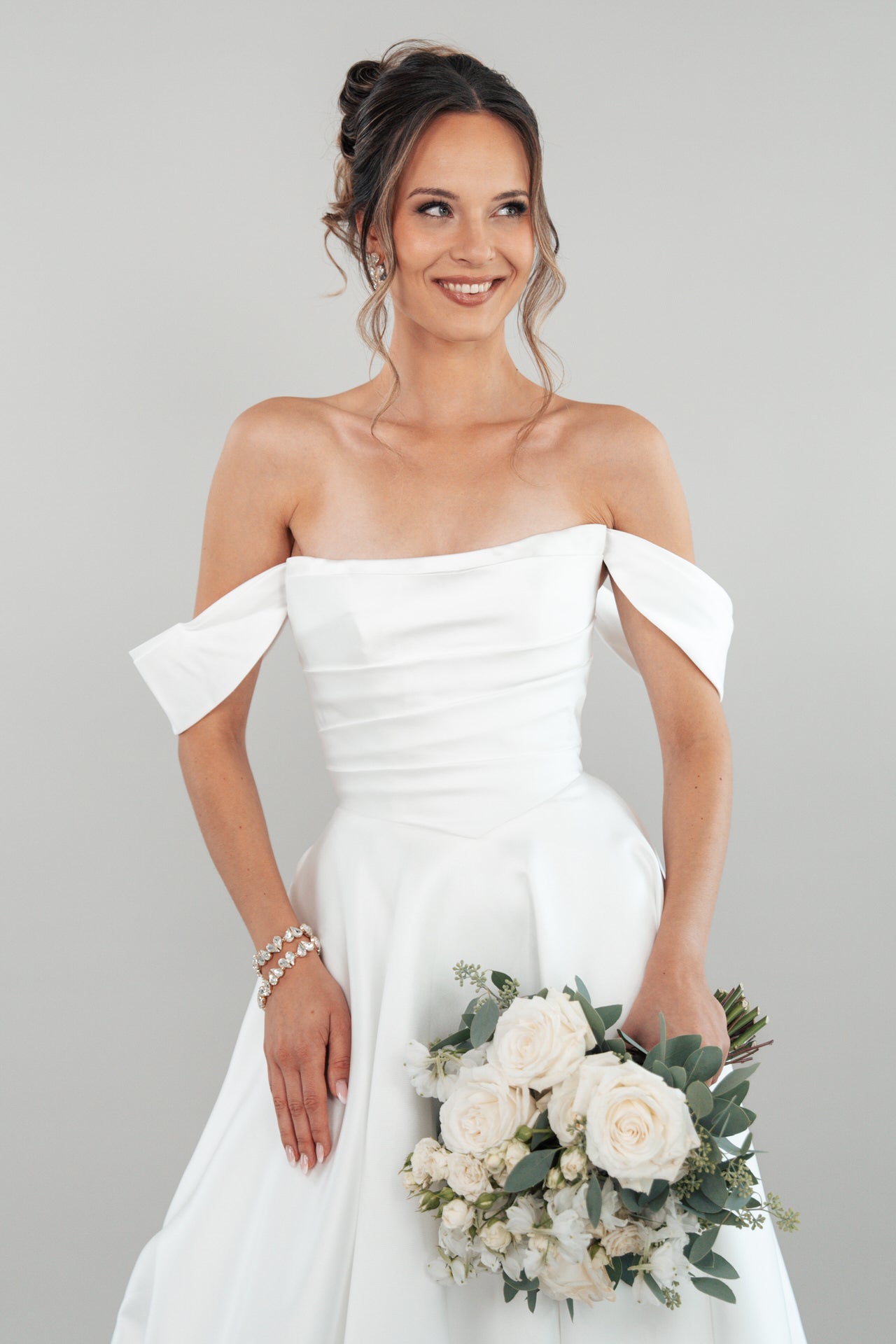 Eine lächelnde Frau modelliert das schulterfreie weiße Hochzeitskleid S26-B01 und hält einen Strauß weißer Rosen und Grünzeug vor einem schlichten hellgrauen Hintergrund.