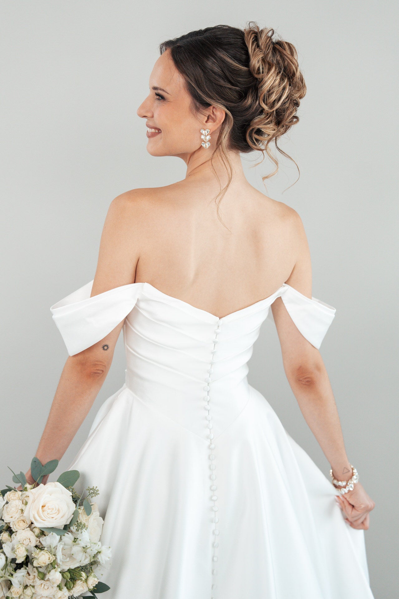 Eine Braut, die das schulterfreie weiße Brautkleid S26-B01 trägt, steht mit einem Strauß weißer und cremefarbener Blumen in der Hand und trägt ihr Haar zu einer eleganten Hochsteckfrisur und Perlenohrringen.