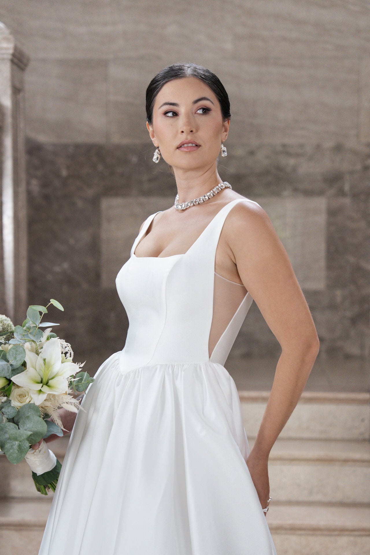 Die Braut trägt das weiße, ärmellose Kleid S26-B02, hält einen Strauß weißer und grüner Blumen in der Hand, trägt Kronleuchter-Ohrringe, eine Halskette und ihr dunkles Haar zurückgestylt, während sie vor Marmorstufen in einer neutralen Umgebung steht.