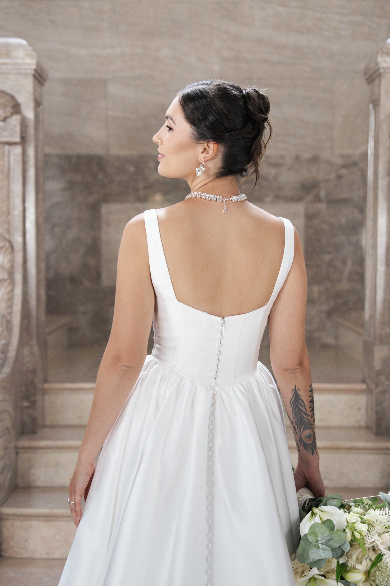 Eine Braut in S26-B02, einem weißen ärmellosen Kleid, steht mit dem Rücken zur Kamera auf steinernen Stufen und hält einen Blumenstrauß. Sie hat eine Tätowierung am rechten Arm, eine elegante Halskette und ihr dunkles Haar ist zu einer Hochsteckfrisur frisiert.
