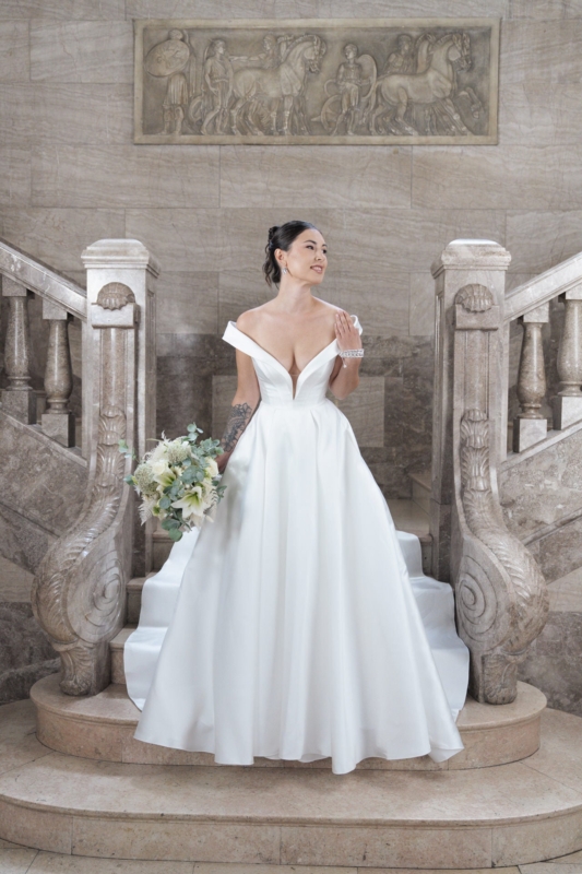 Eine Braut in dem schulterfreien weißen Kleid S26-B03 hält einen Blumenstrauß, während sie auf einer großen Steintreppe steht, über der sich ein verziertes Geländer und geschnitzte Kunstwerke befinden.