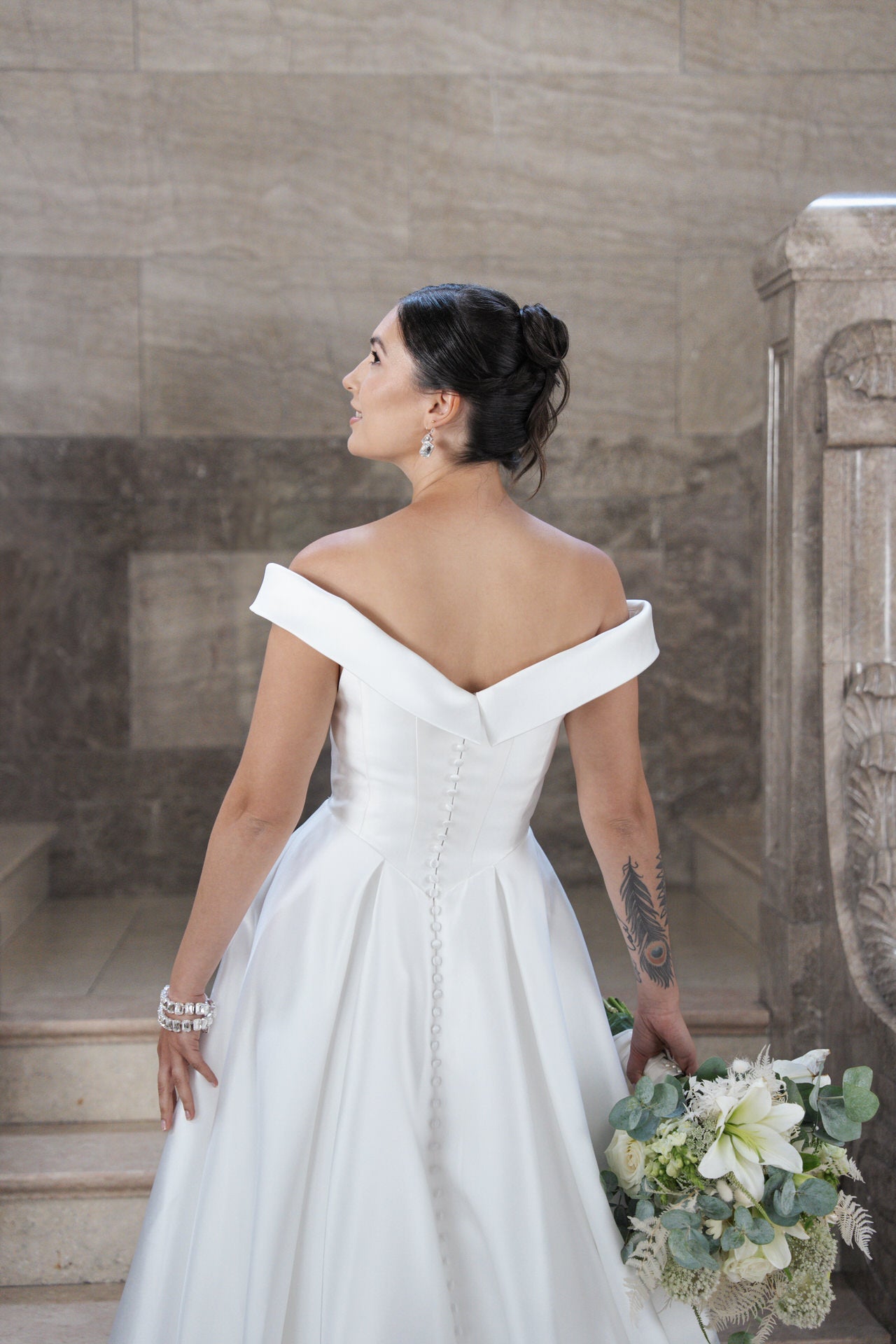In dem schulterfreien weißen Kleid S26-B03 steht eine Braut mit dunklem Dutt und sichtbarem Armtattoo auf einer Treppe und hält einen Strauß mit weißen Blumen und Grünzeug in der Hand, während sie nach oben blickt.