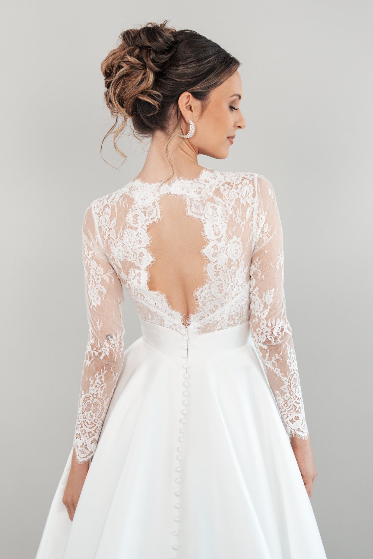 Eine Frau in dem Hochzeitskleid S26-B04 mit Spitzenärmeln und tiefem Rückenausschnitt steht mit einer eleganten Hochsteckfrisur vor einem schlichten hellgrauen Hintergrund.