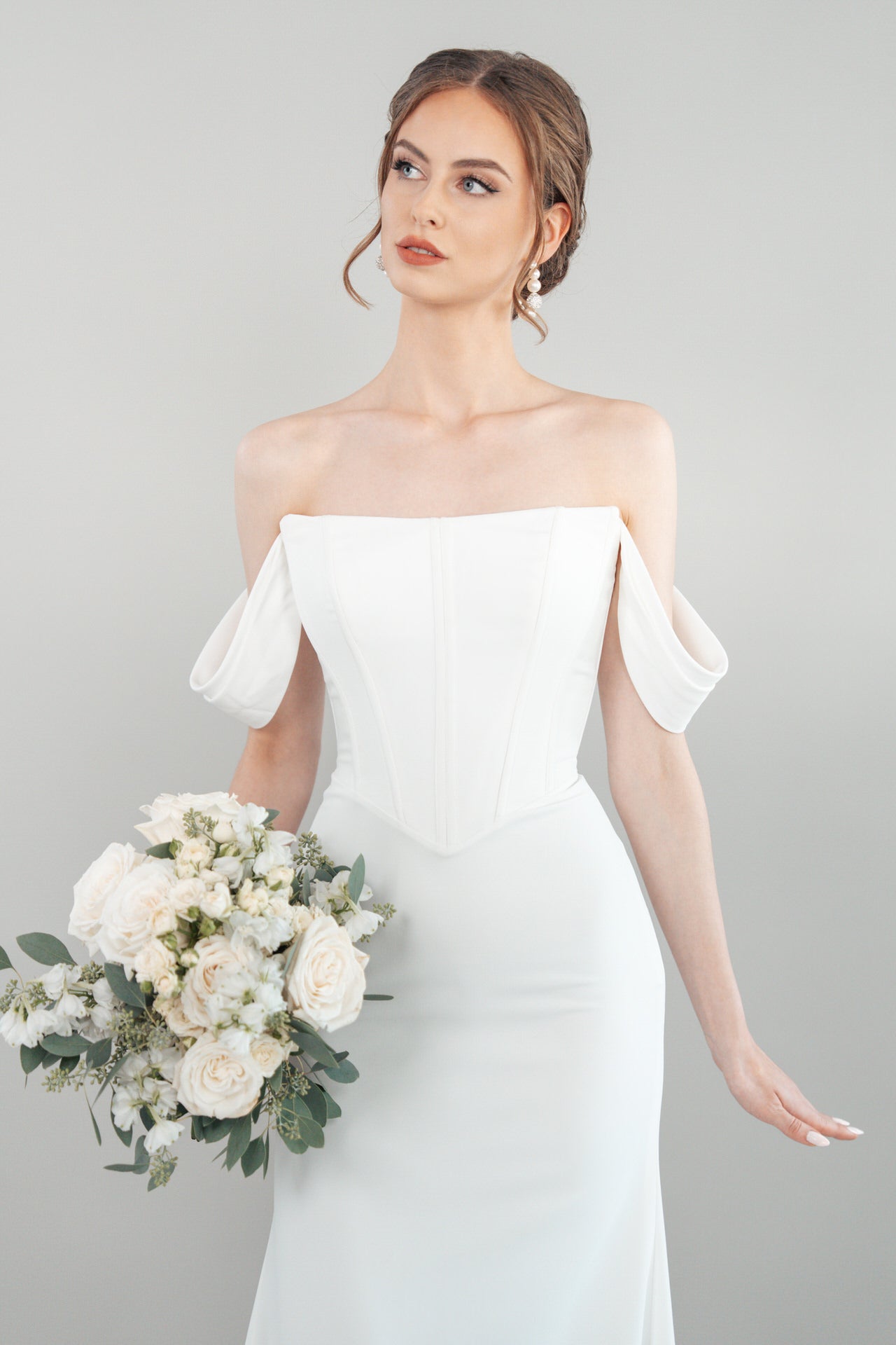 Eine Frau modelliert das schulterfreie weiße Hochzeitskleid S26-F01 und hält einen Strauß weißer Blumen und Grünzeug vor einem schlichten hellgrauen Hintergrund. Ihr Haar ist hochgesteckt, während sie leicht nach oben blickt.