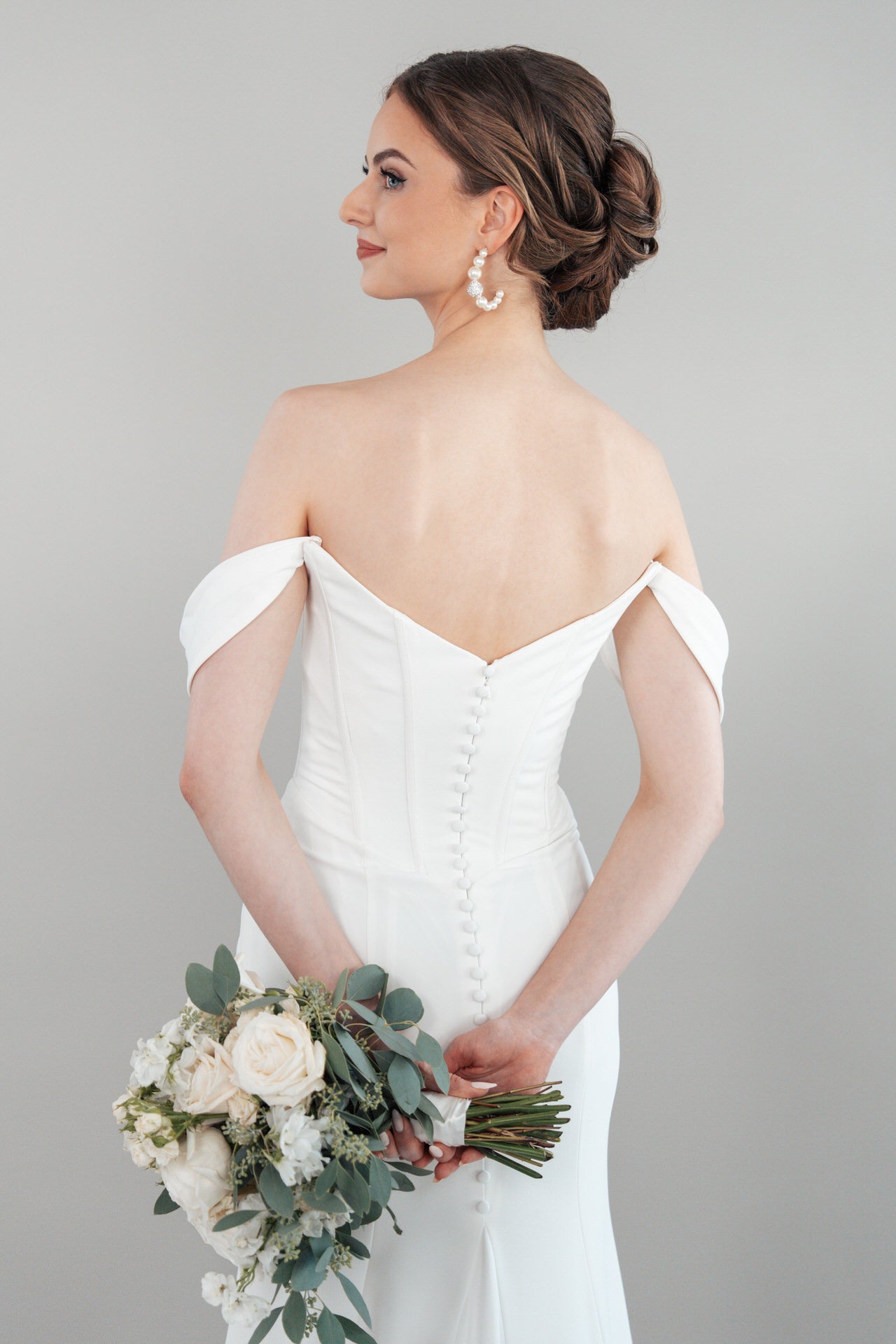 Eine Frau, die das schulterfreie weiße Hochzeitskleid S26-F01 trägt, steht mit dem Rücken zugewandt und hält einen Strauß mit weißen Blumen und Grünzeug in der Hand. Ihr Haar ist elegant gestylt und sie trägt Perlenohrringe.