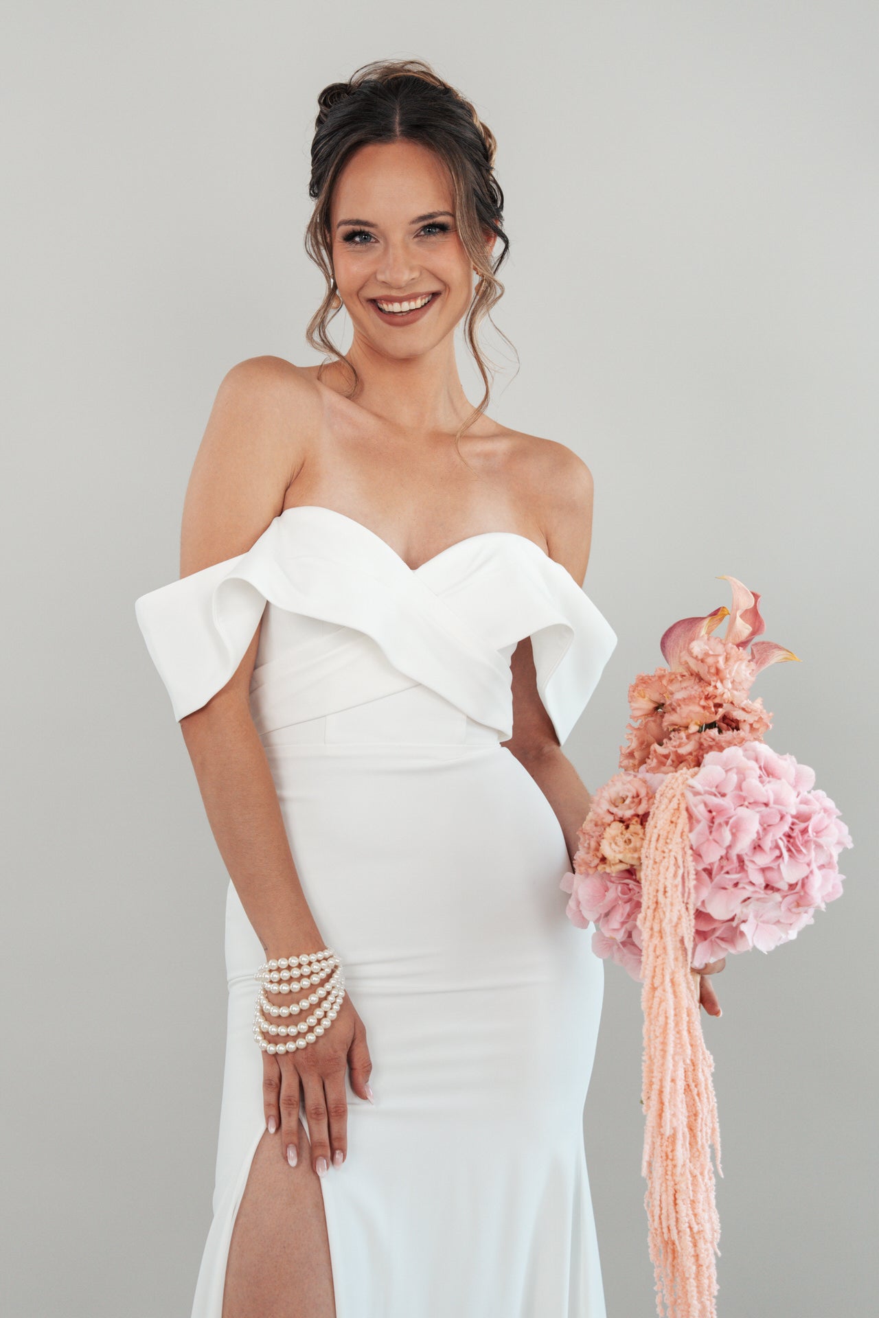 Eine lächelnde Frau im schulterfreien weißen Kleid S26-F02 hält rosa Blumen, ihr gewelltes Haar ist hochgesteckt und sie trägt ein Perlenarmband am Handgelenk. Der Hintergrund ist einfarbig und hell.
