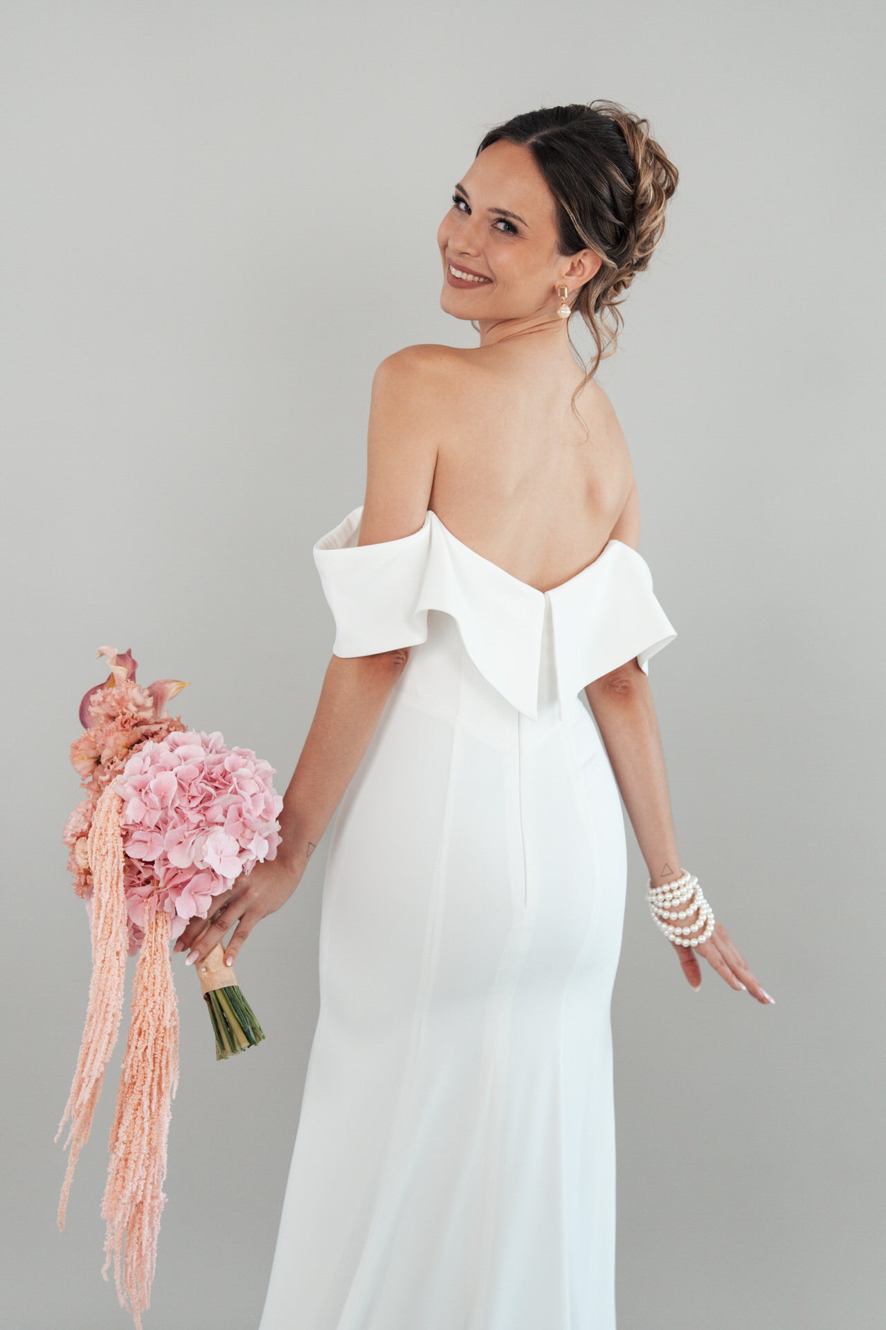 Eine Frau in dem schulterfreien weißen Kleid S26-F02 lächelt über ihre Schulter und hält rosa Blumen in der Hand. Ihr Haar ist hochgesteckt, sie trägt ein Perlenarmband und steht vor einem schlichten hellen Hintergrund.