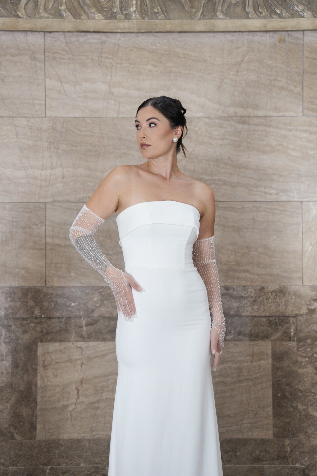 Eine Frau in dem trägerlosen weißen Kleid S26-F03 und durchsichtigen, perlenbesetzten Handschuhen steht an einer beigen und braunen Marmorwand und blickt mit der Hand in der Hüfte zur Seite. Ihr dunkles Haar ist zu einer eleganten Hochsteckfrisur gestylt.