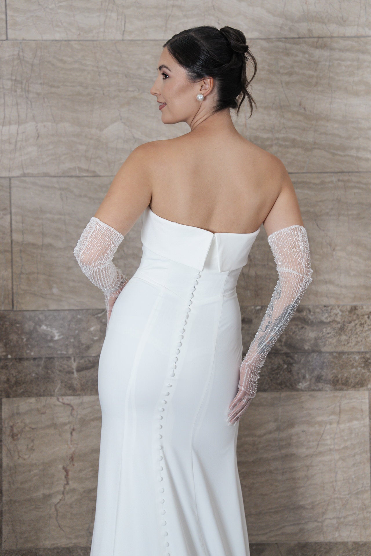 Eine Frau modelliert das trägerlose weiße Kleid S26-F03 mit Perlenknöpfen am Rücken, durchsichtigen Perlenhandschuhen und einer eleganten Hochsteckfrisur und posiert vor einer Steinmauer.