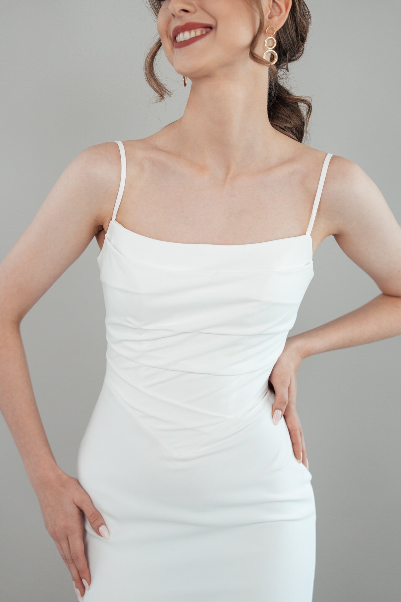 Eine Frau in einem weißen Kleid mit dünnen Trägern S26-F04 lächelt mit den Händen in den Hüften. Sie hat gewelltes braunes Haar in einem Pferdeschwanz und trägt große goldene geometrische Ohrringe. Der Hintergrund ist einfarbig hellgrau.