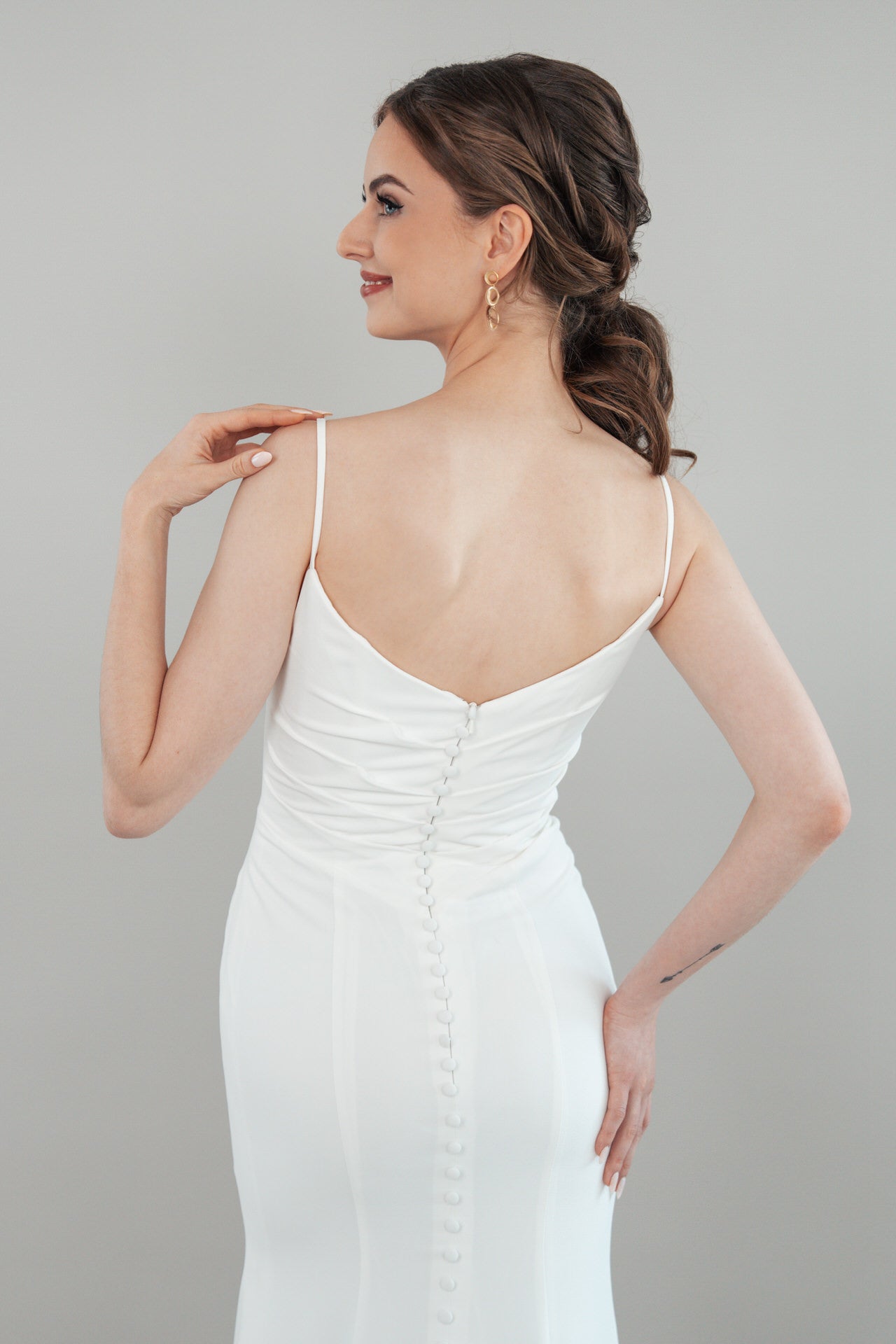 Eine Frau, die S26-F04, ein weißes ärmelloses Kleid mit Knopfleiste, trägt, steht zur Seite gedreht und lächelt. Ihr Haar ist in lockeren Wellen nach hinten gestylt, und sie trägt goldene Ohrringe. Der Hintergrund ist einfarbig hellgrau.