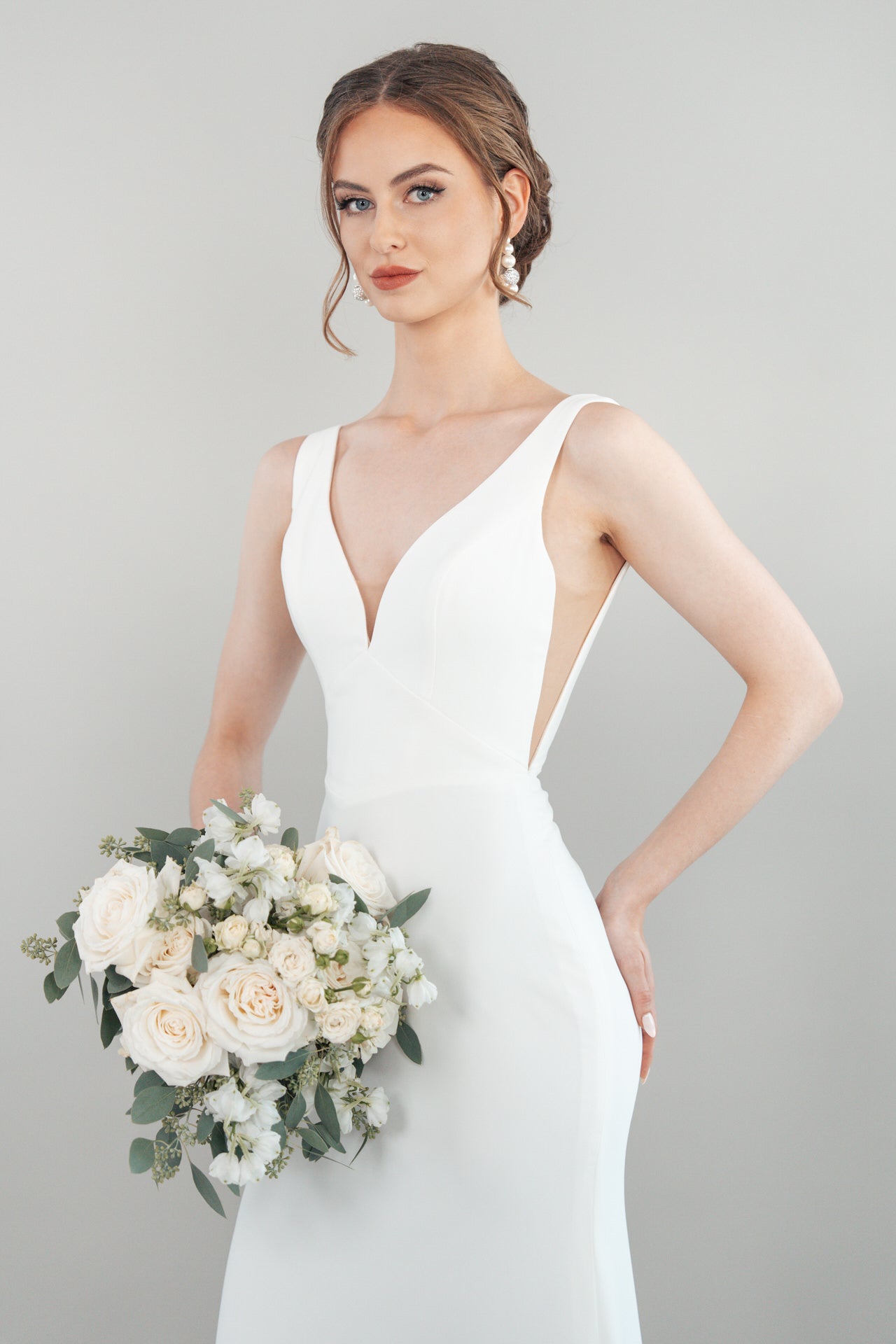 Eine Frau, die das weiße ärmellose Hochzeitskleid S26-F05 trägt, hält einen Strauß weißer Blumen und Grünzeug vor einem schlichten hellgrauen Hintergrund. Sie hat gestyltes braunes Haar, auffällige Ohrringe und einen gelassenen Gesichtsausdruck.