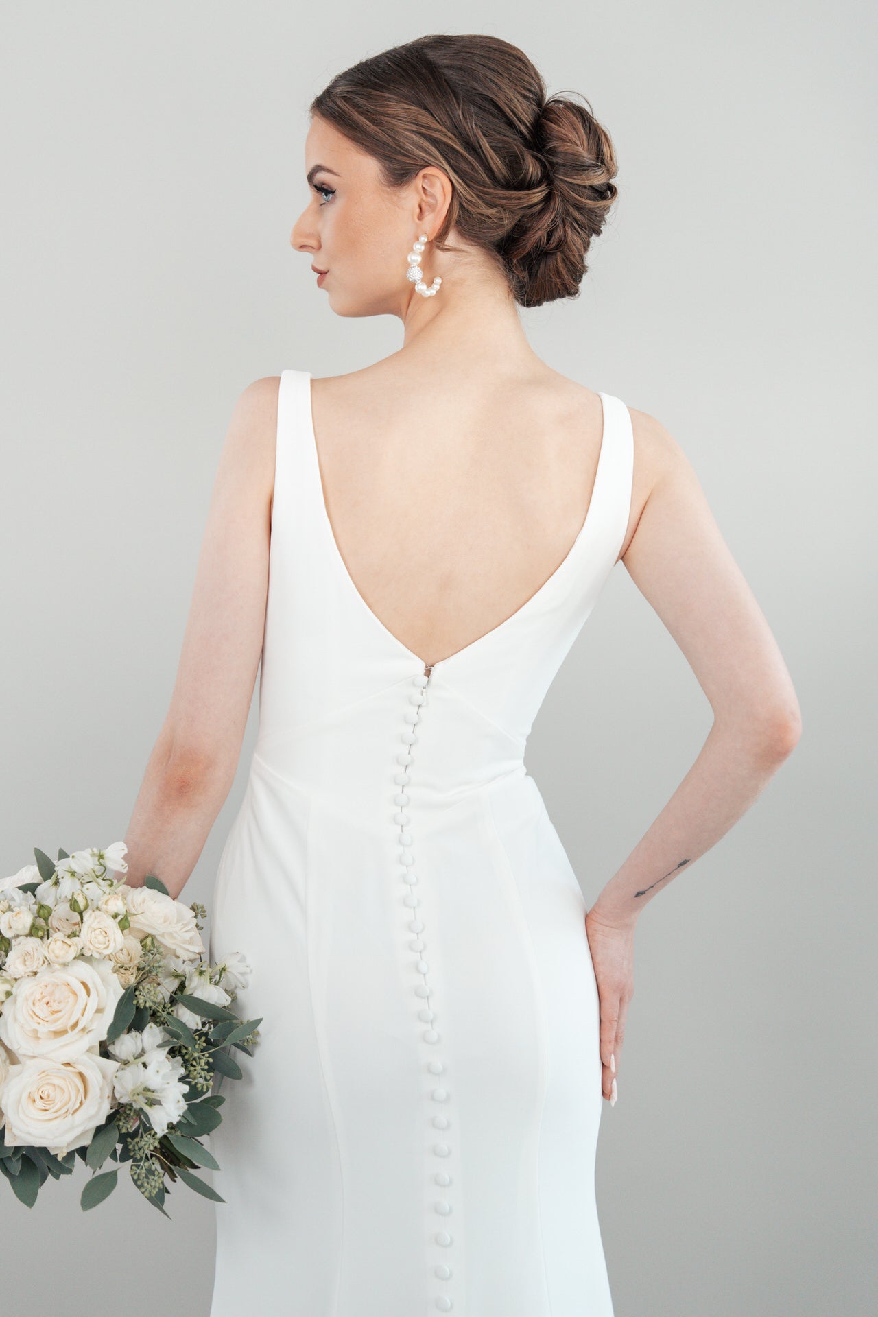 Eine Braut in Produkt S26-F05, einem weißen ärmellosen Kleid mit tiefem V-Ausschnitt und Knopfleiste, hält einen Strauß aus weißen Rosen und Grünzeug. Ihr Haar ist zu einer eleganten Hochsteckfrisur frisiert, während sie zur Seite blickt.