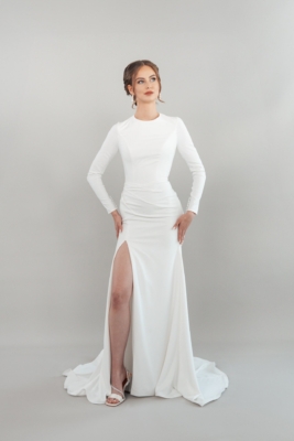 Eine Frau posiert selbstbewusst mit den Händen in den Hüften in S26-F06 - einem taillierten, langärmeligen weißen Kleid mit hohem Schlitz - vor einem schlichten, hellgrauen Hintergrund.