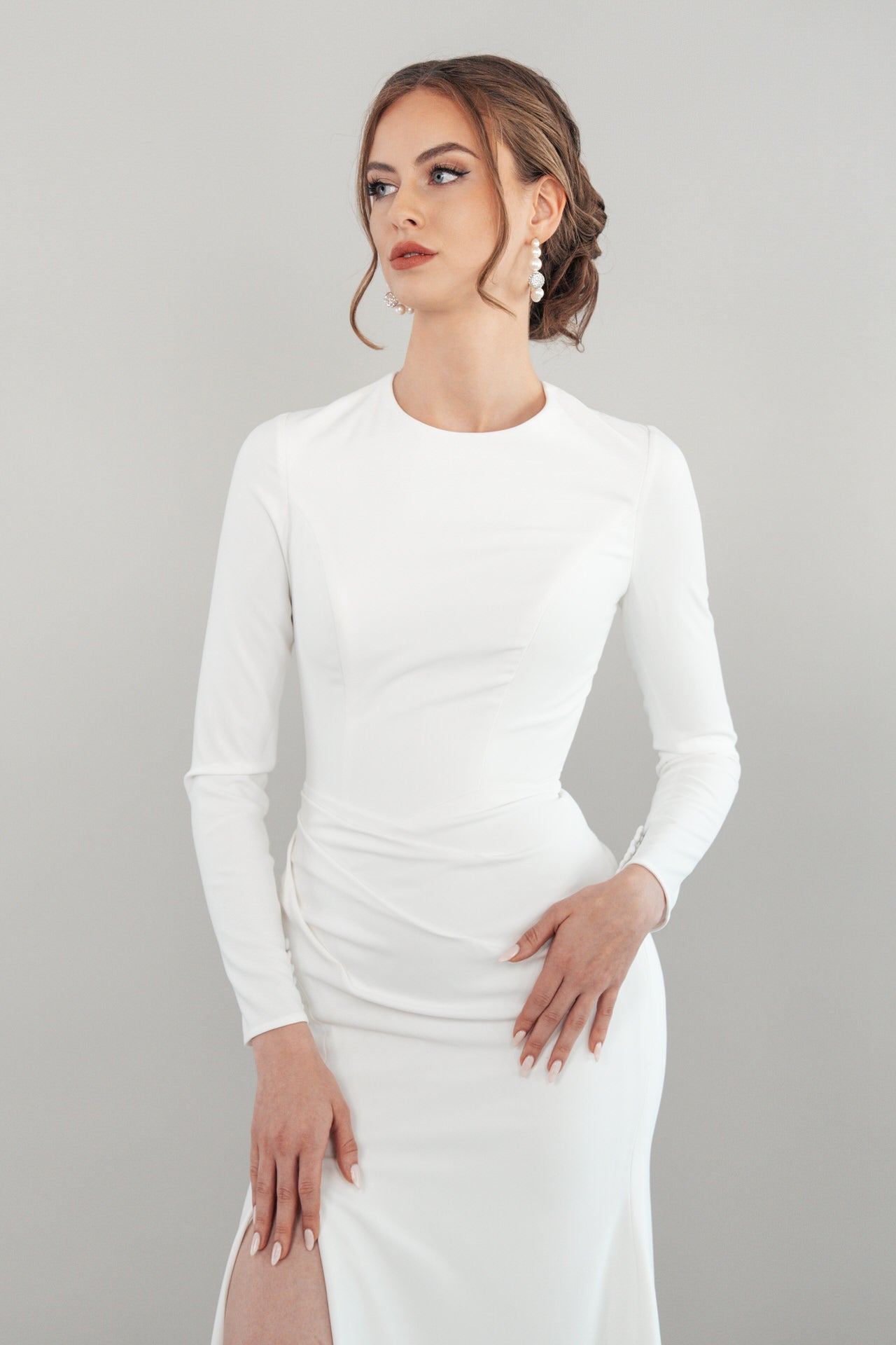 Eine Frau, die das langärmelige weiße Kleid S26-F06 mit taillierter Passform trägt, posiert selbstbewusst vor einem hellgrauen Hintergrund. Ihr Haar ist zu einer eleganten Hochsteckfrisur frisiert und mit Ohrringen geschmückt, wobei sie leicht zur Seite schaut.