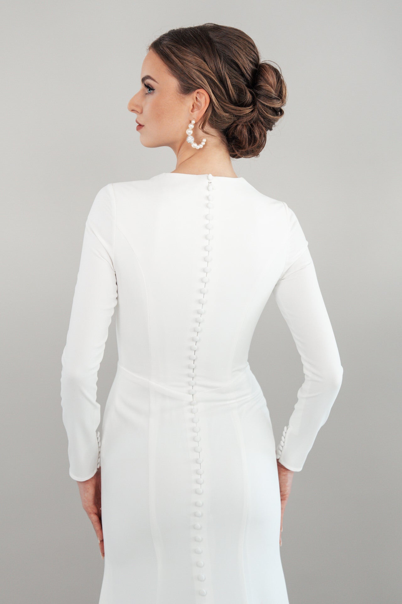 In S26-F06, einem langärmeligen, taillierten weißen Kleid mit Knöpfen am Rücken, steht eine Frau mit abgewandtem Blick und gedrehtem Kopf. Ihr Haar ist zu einer eleganten Hochsteckfrisur gestylt, die durch auffällige Perlenohrringe ergänzt wird.