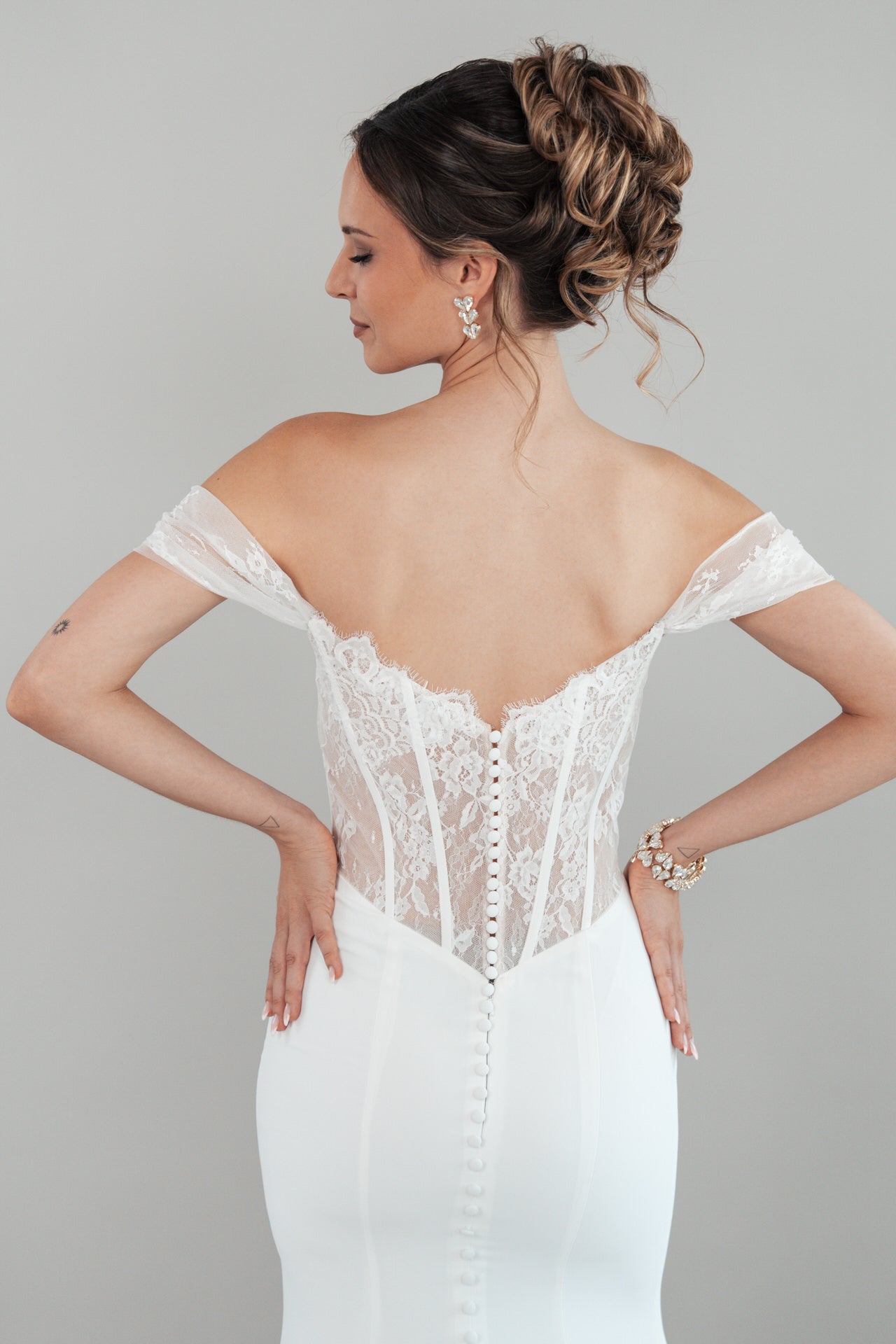 Eine Frau mit einer eleganten Hochsteckfrisur modelliert das S26-F07, ein schulterfreies, weißes Hochzeitskleid mit Spitzenrücken. Sie posiert mit dem Rücken zur Kamera und den Händen in den Hüften vor einem schlichten Hintergrund.