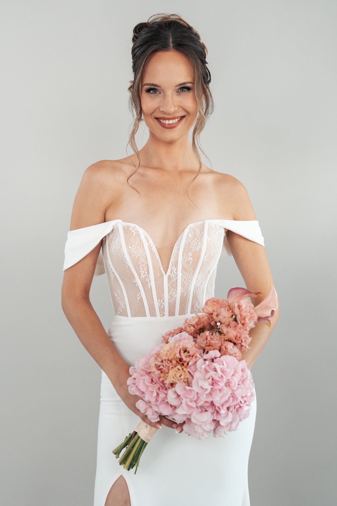 Eine lächelnde Frau trägt das schulterfreie weiße Hochzeitskleid S26-F08 und hält einen Strauß rosa Blumen. Ihr Haar ist hochgesteckt, lose Strähnen umrahmen ihr Gesicht, und sie steht vor einem schlichten hellgrauen Hintergrund.