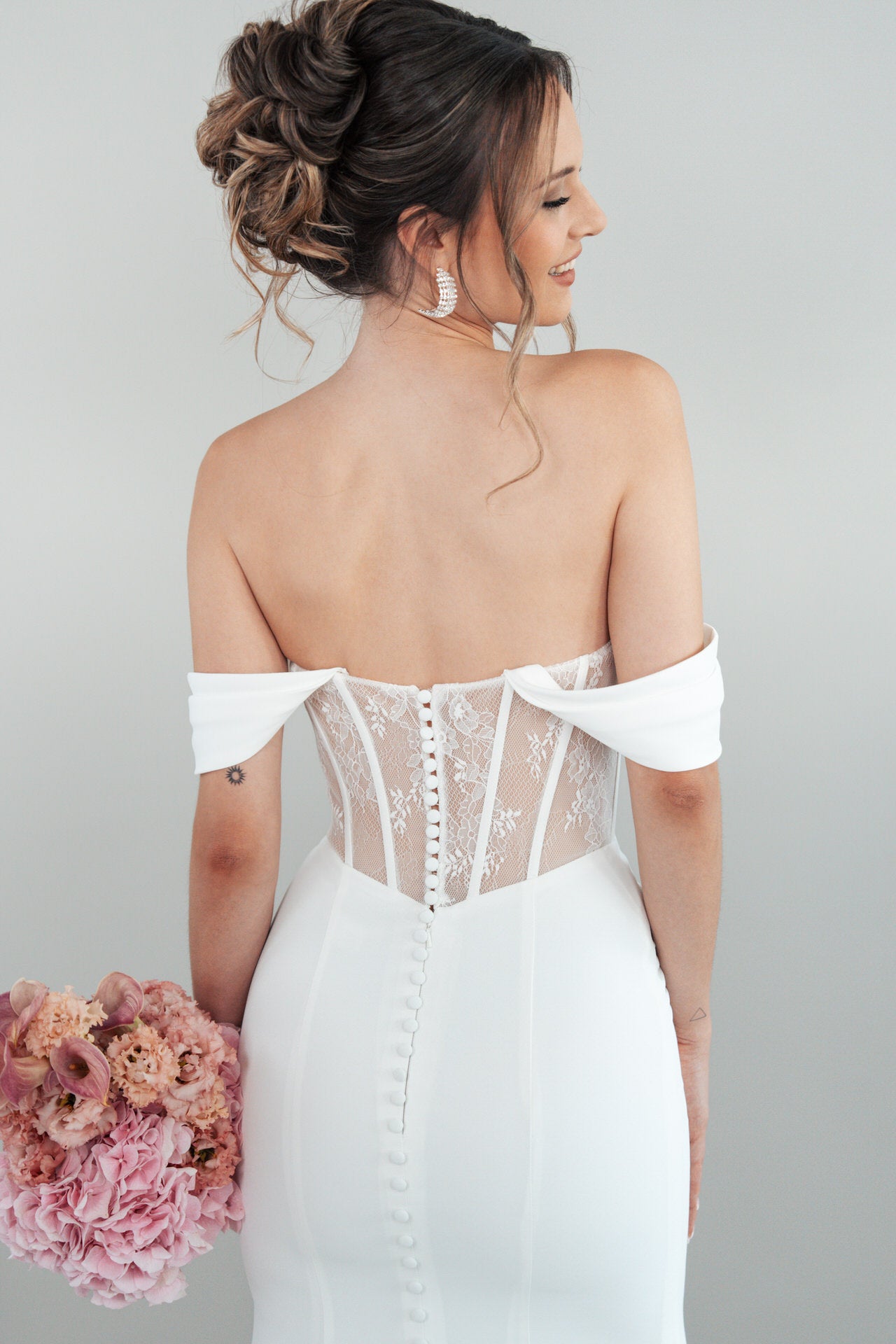 In dem schulterfreien weißen Hochzeitskleid S26-F08 mit Spitze und Knöpfen hält eine Frau einen rosafarbenen Blumenstrauß und blickt mit einem sanften Lächeln über ihre Schulter.