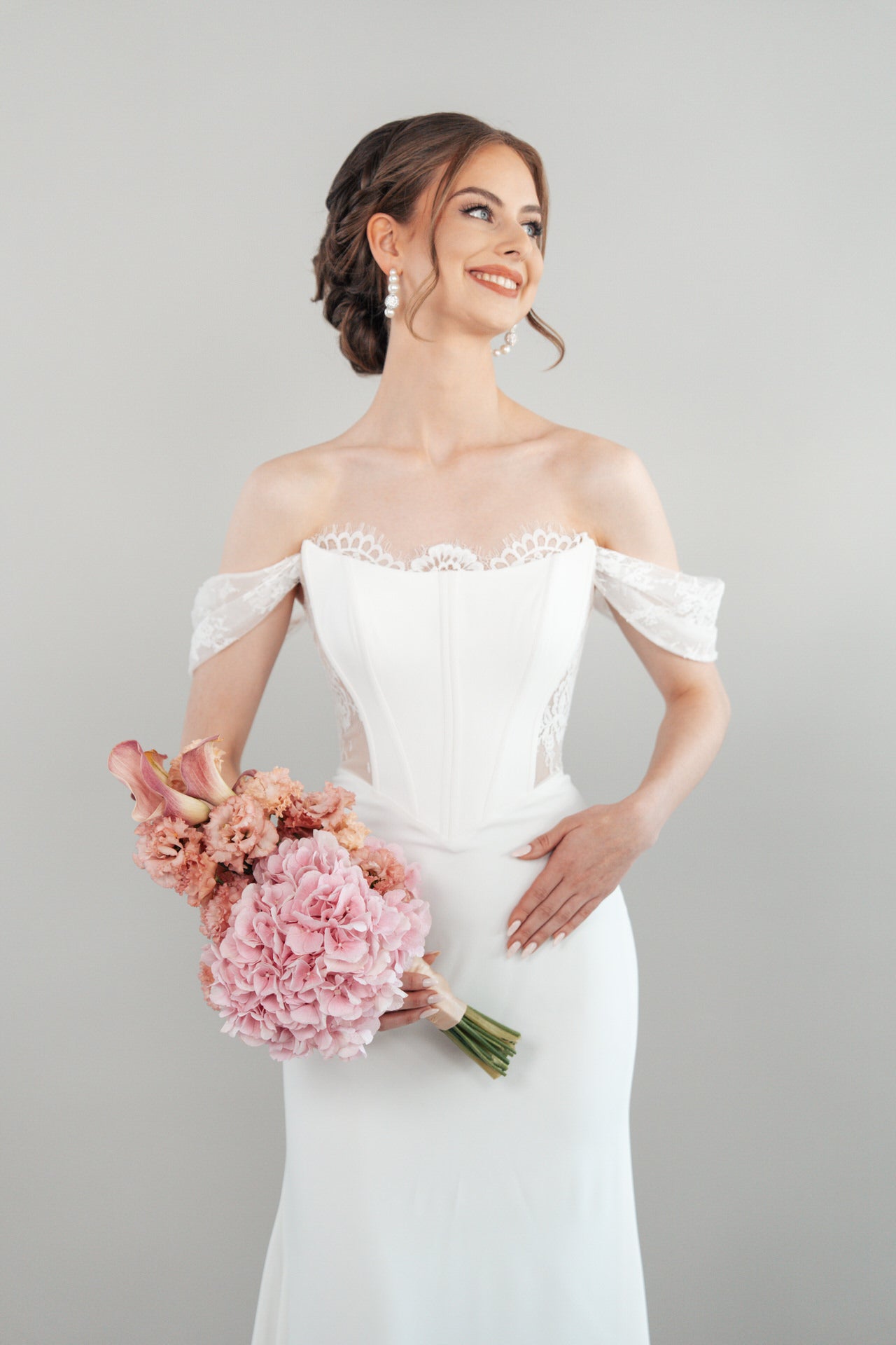 Eine lächelnde Braut im schulterfreien weißen Hochzeitskleid S26-F09 hält einen Strauß rosafarbener Blumen und blickt zur Seite vor einem schlichten hellen Hintergrund.