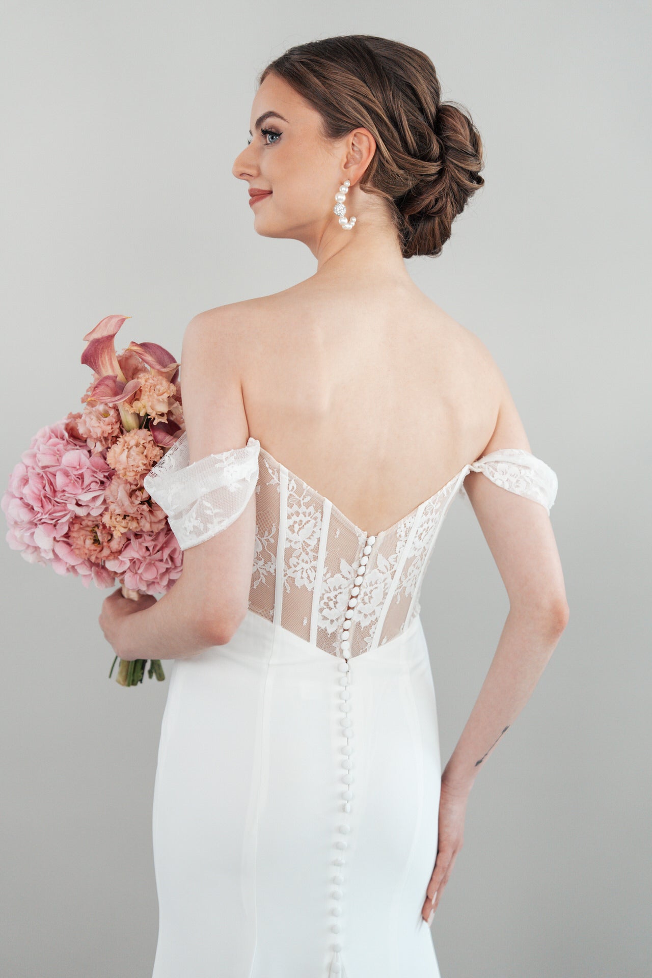 Vor einem schlichten Hintergrund steht eine Braut im schulterfreien weißen Brautkleid S26-F09 mit Spitzenverzierung am Rücken, die Haare zu einer eleganten Hochsteckfrisur frisiert, mit Perlenohrringen und einem Strauß rosa Blumen in der Hand.
