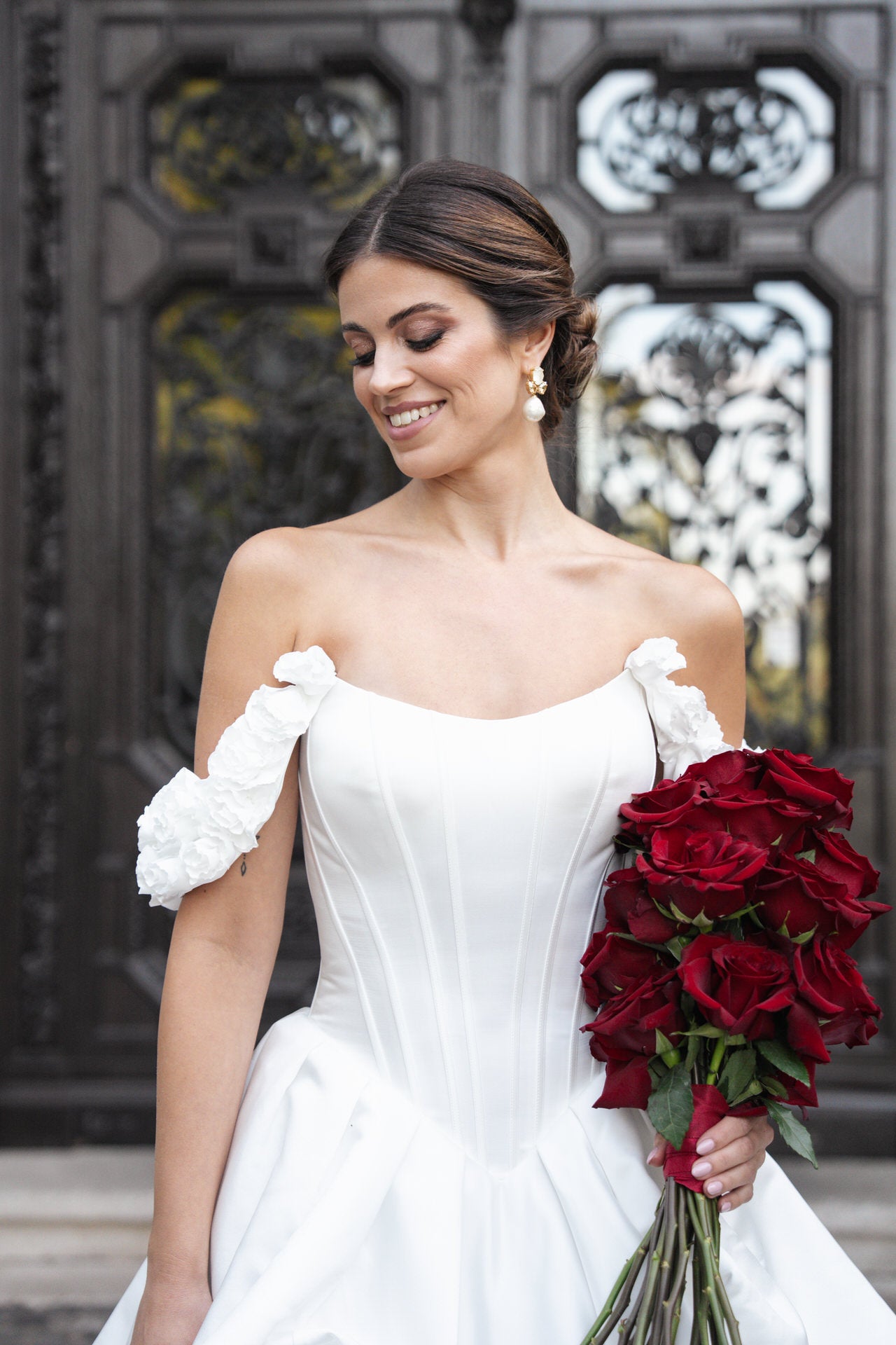 hochzeitsrausch Brautmoden - Elegantes Brautkleid mit schulterfreien Ärmeln und Rosenstrauß vor kunstvoller Tür