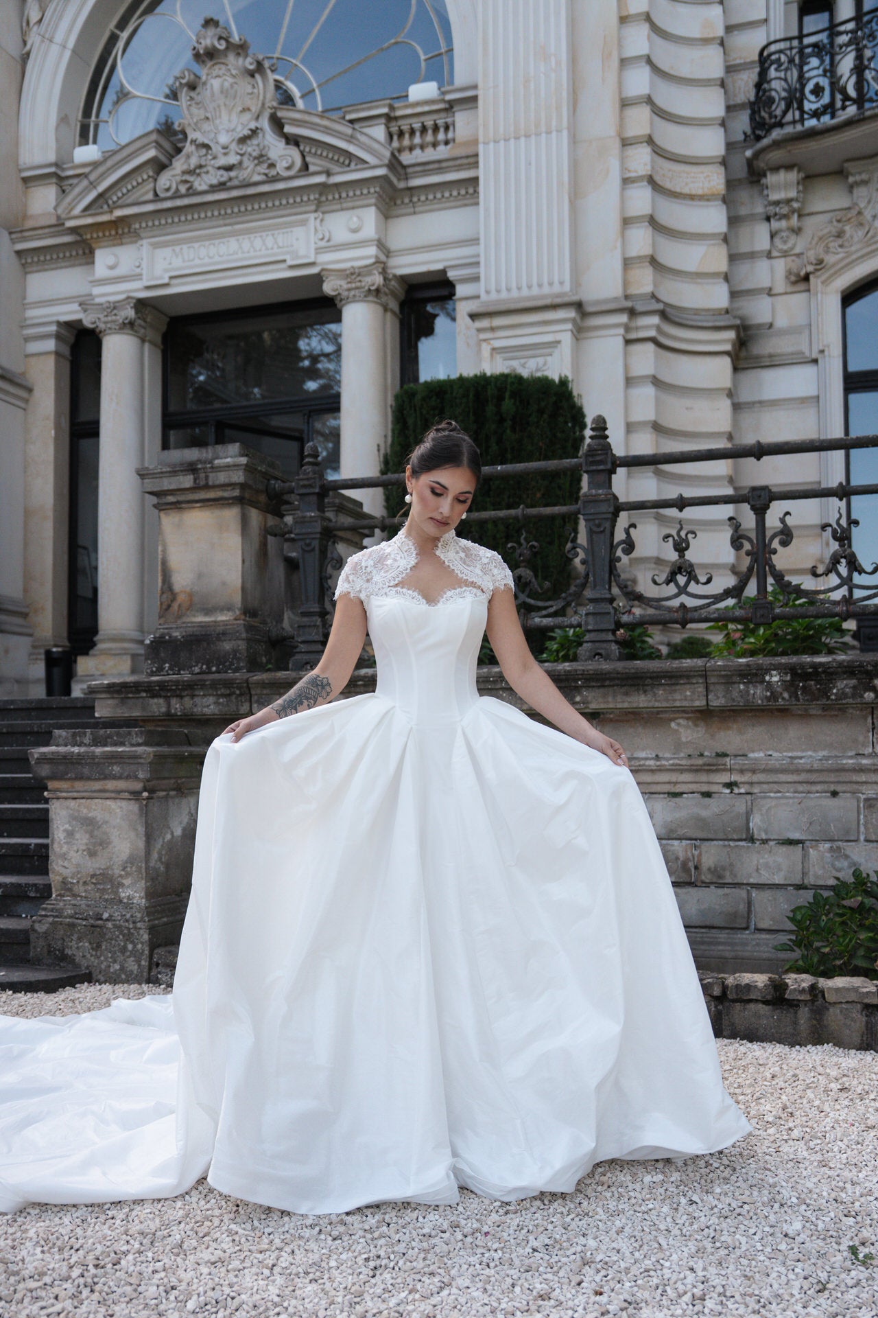 Braut in elegantem, weißem Kleid mit Spitzenapplikationen, vor historischer Gebäude-Fassade, romantische und edle Atmosphäre.